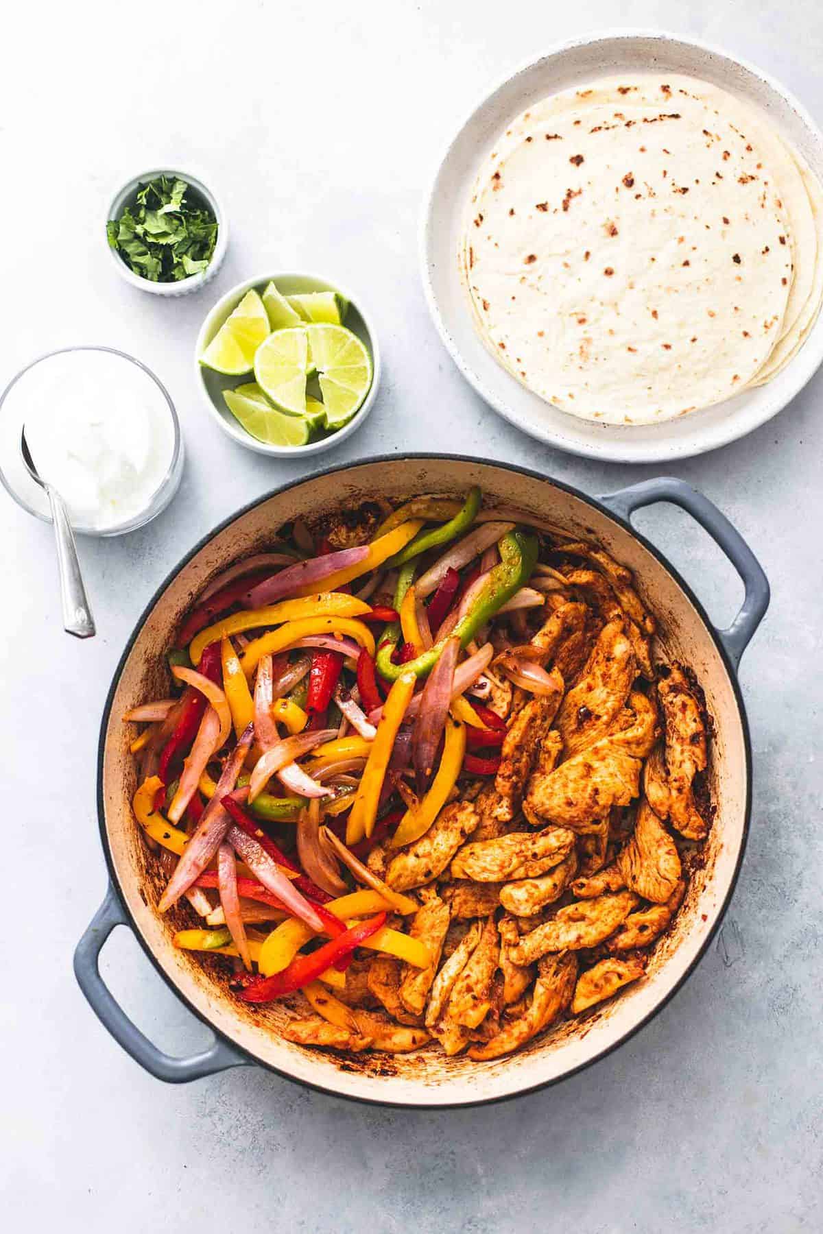 top view of chicken fajitas skillet with bowls of lime slices, cilantro and sour cream and a plate of tortillas on the side.