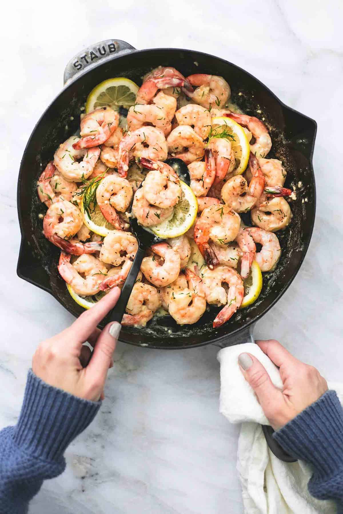 top view of a hand holding a handle of  a pan of creamy lemon dill shrimp with another hand scooping some shrimp with a serving spoon.