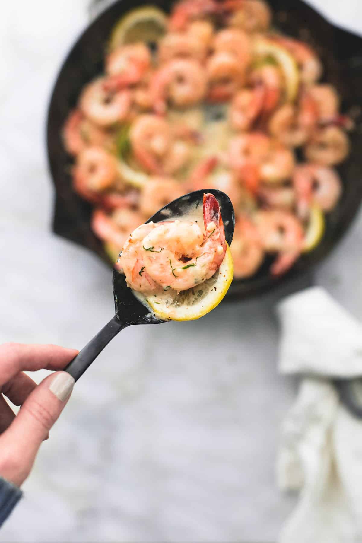 a hand holding creamy lemon dill shrimp in a serving spoon above a pan of more shrimp.