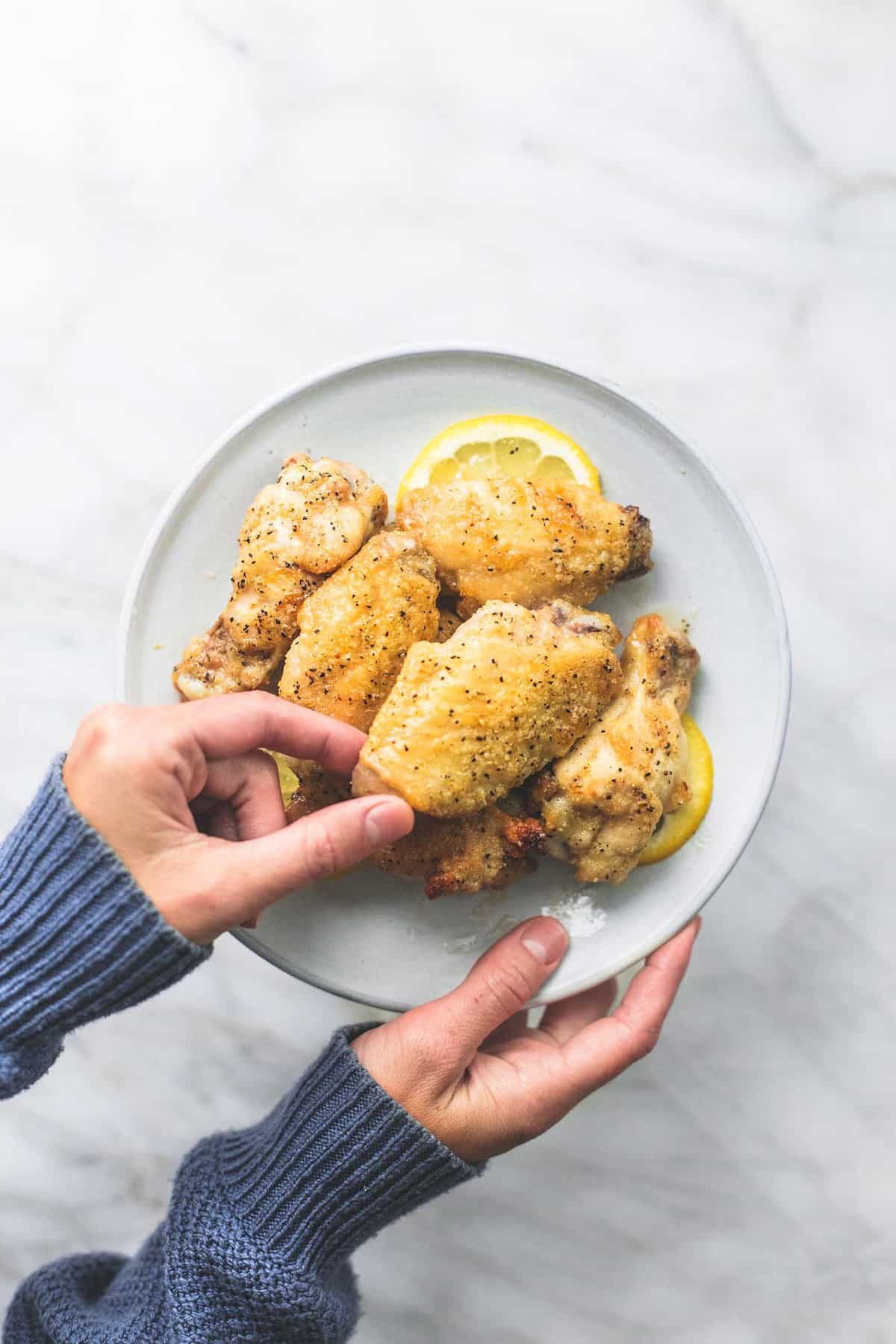 top view of a hand holding a plate of baked lemon pepper chicken wings with another hand grabbing a wing.