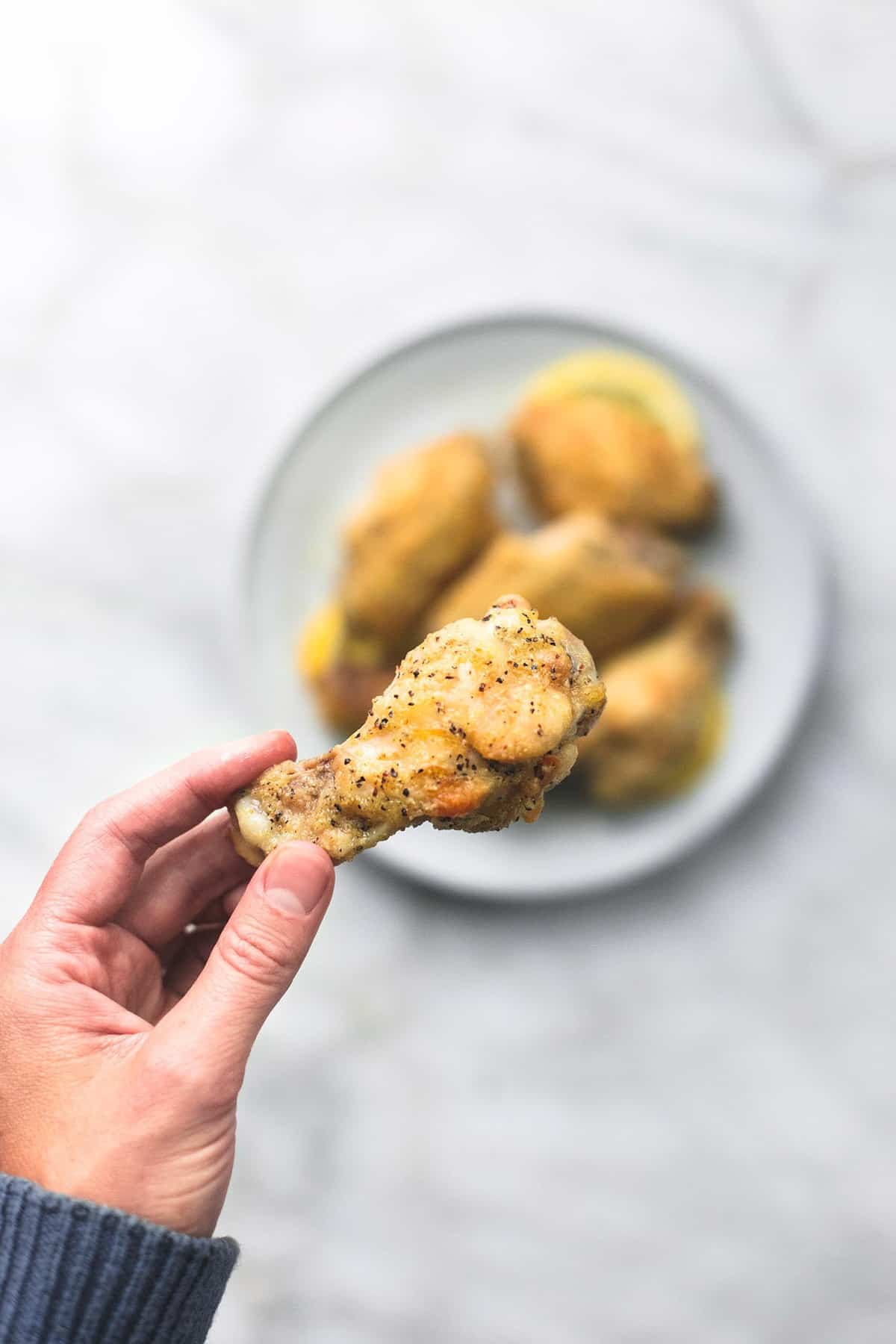 a hand holding a baked lemon pepper chicken wing above a plate of more wings.