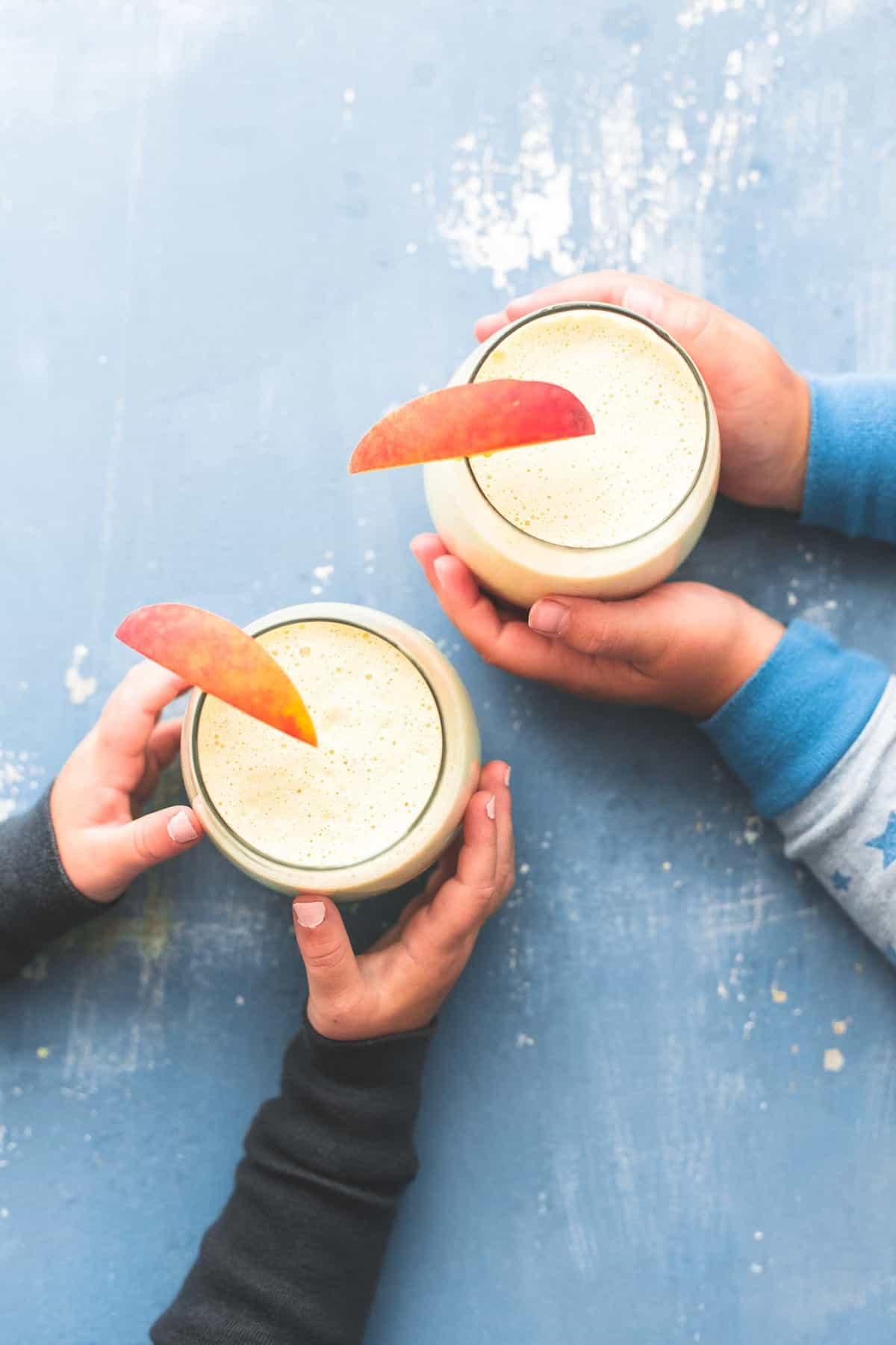 top view of two pairs of child's hands each grabbing a glass of peaches n cream moon milk with a peach slice on top.