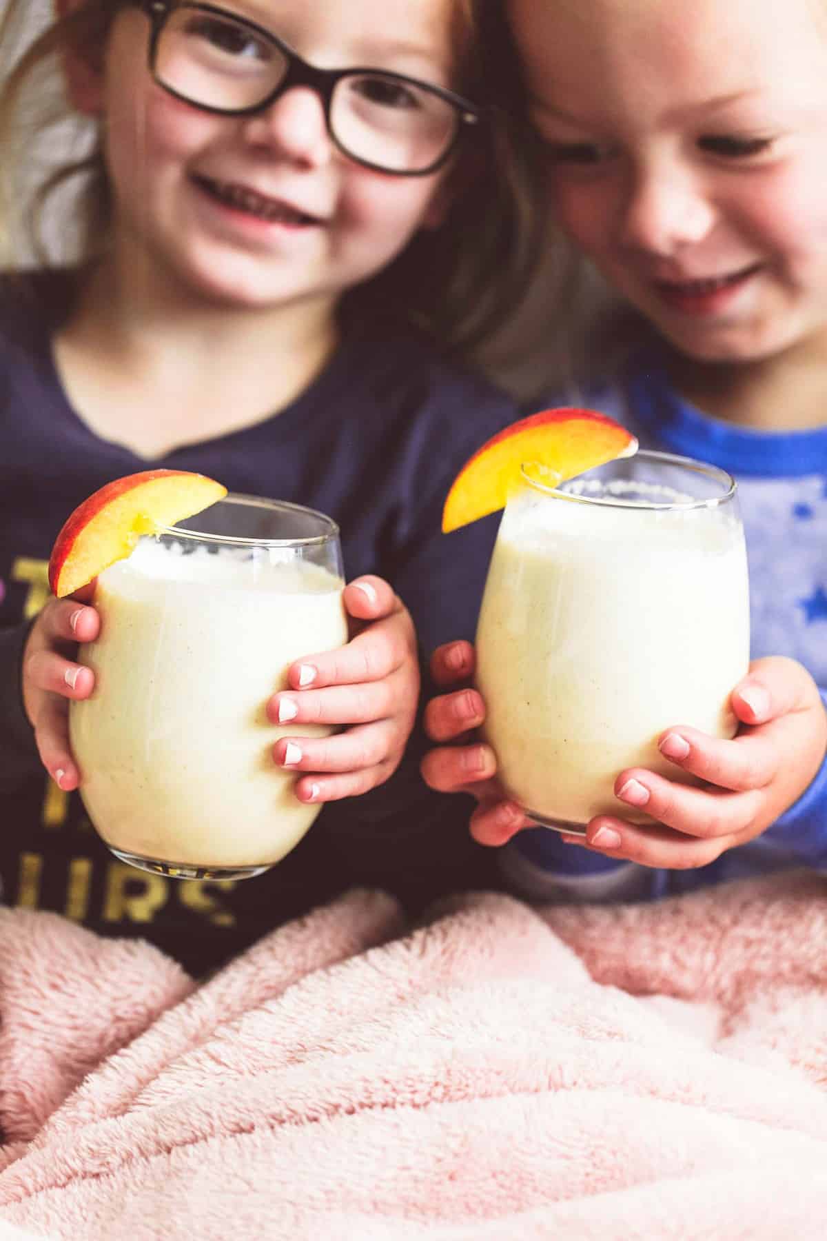 a little girl and boy holding a glass each of peaches n cream moon milk with a peach slice on the top.