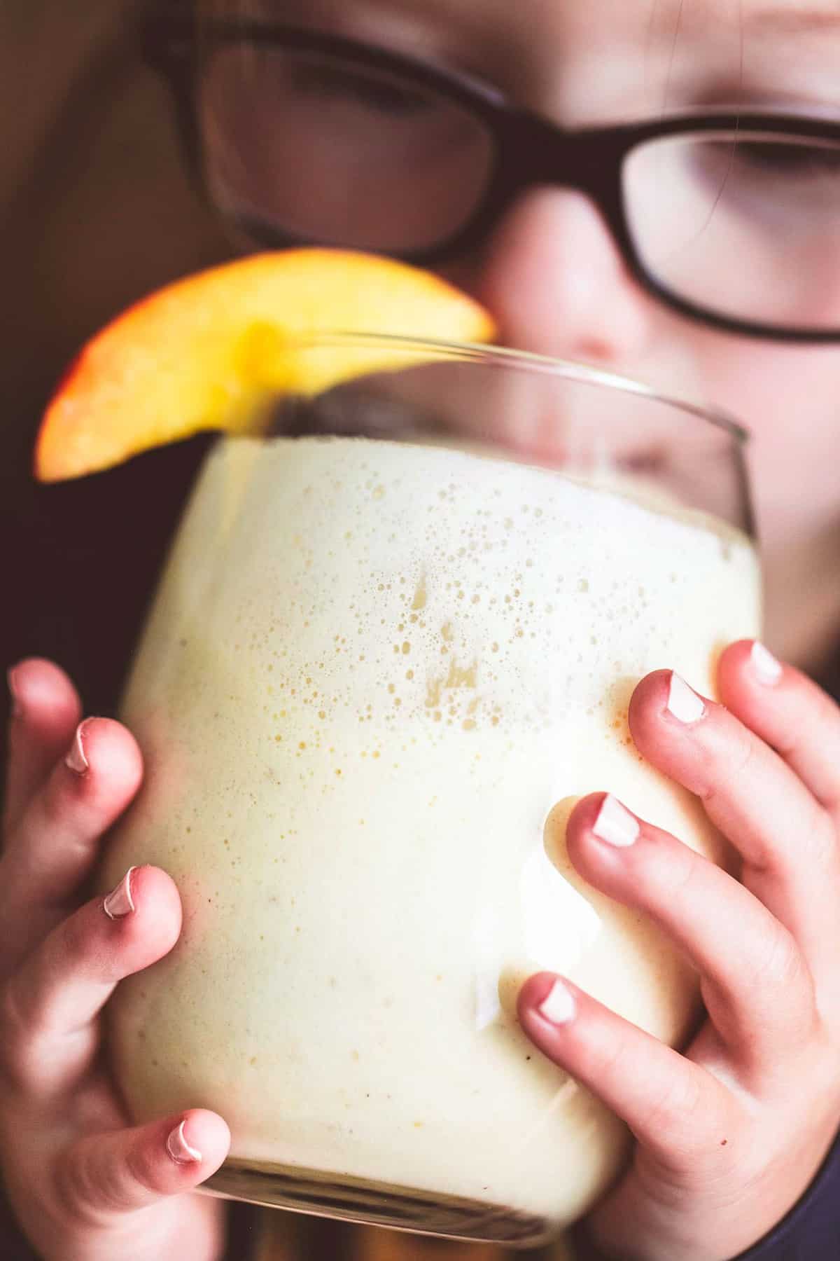 close up of a child drinking peaches n cream moon milk from a glass with a peach slice on top.