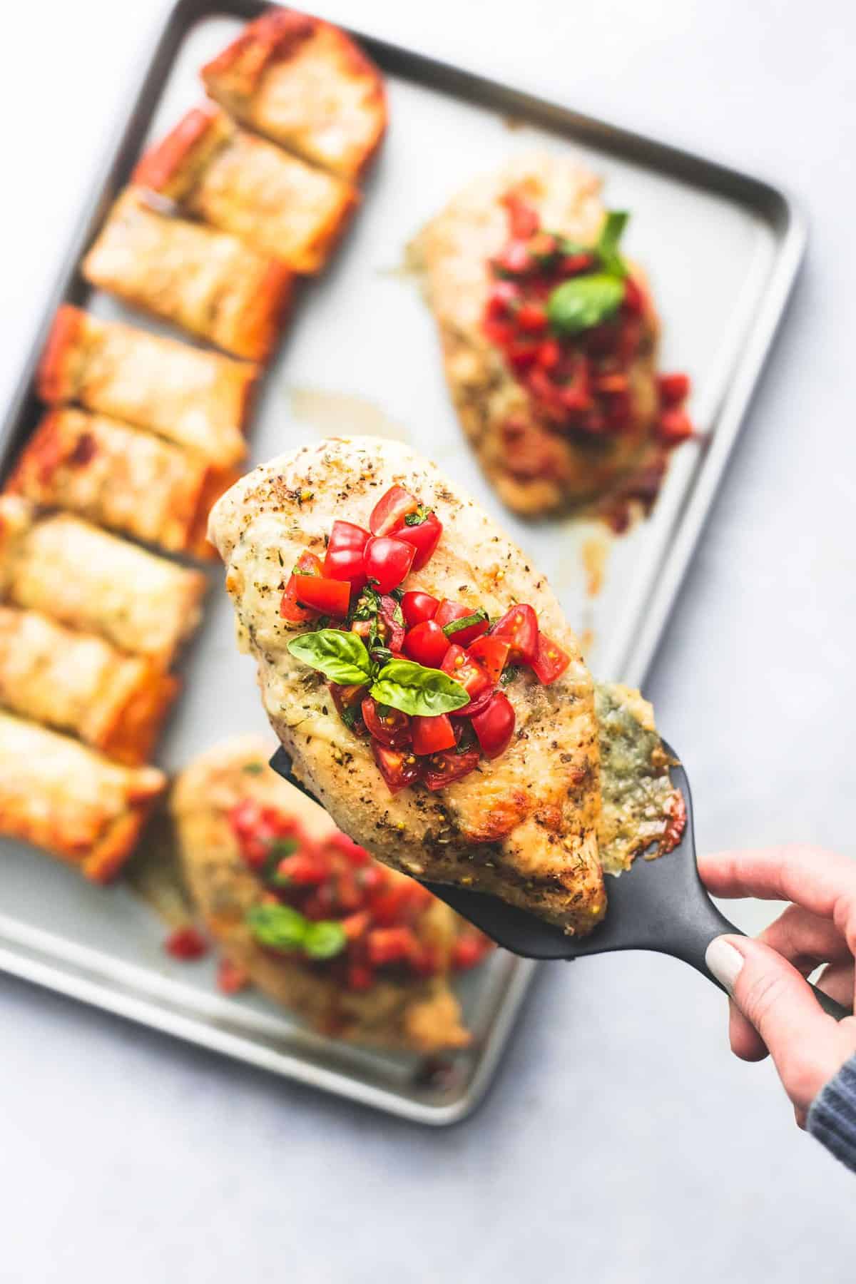 top view of a piece of sheet pan bruschetta chicken being held up above a sheet pan of more chicken and cheesy garlic bread.