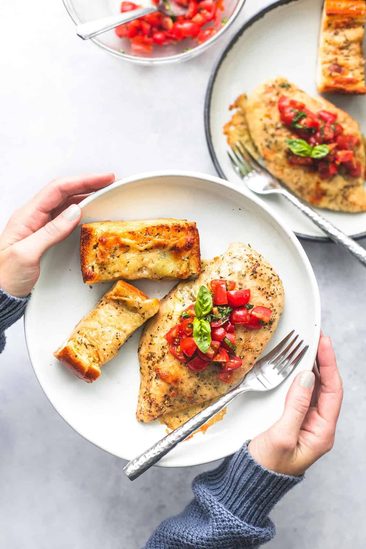 a pair of hands holding a plate with sheet pan bruschetta chicken and cheesy garlic bread and a fork on a it with another plate and a glass bowl of tomatoes on the side.