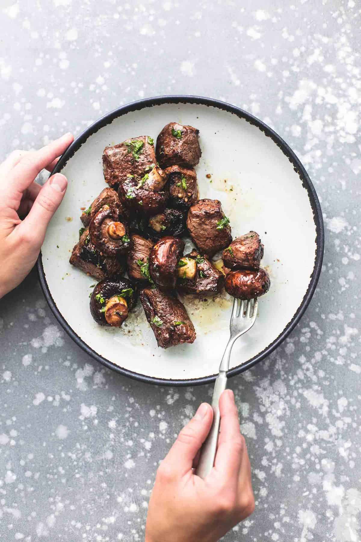 top view of deconstructed grilled steak and mushroom kabobs on a plate with a hand holding the plate and another holding a fork with a piece of the kabobs on it.