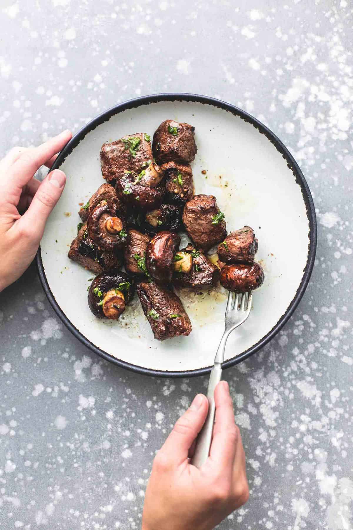 top view of deconstructed grilled steak and mushroom kabobs on a plate with a hand holding the plate and another holding a fork with a piece of the kabobs on it.