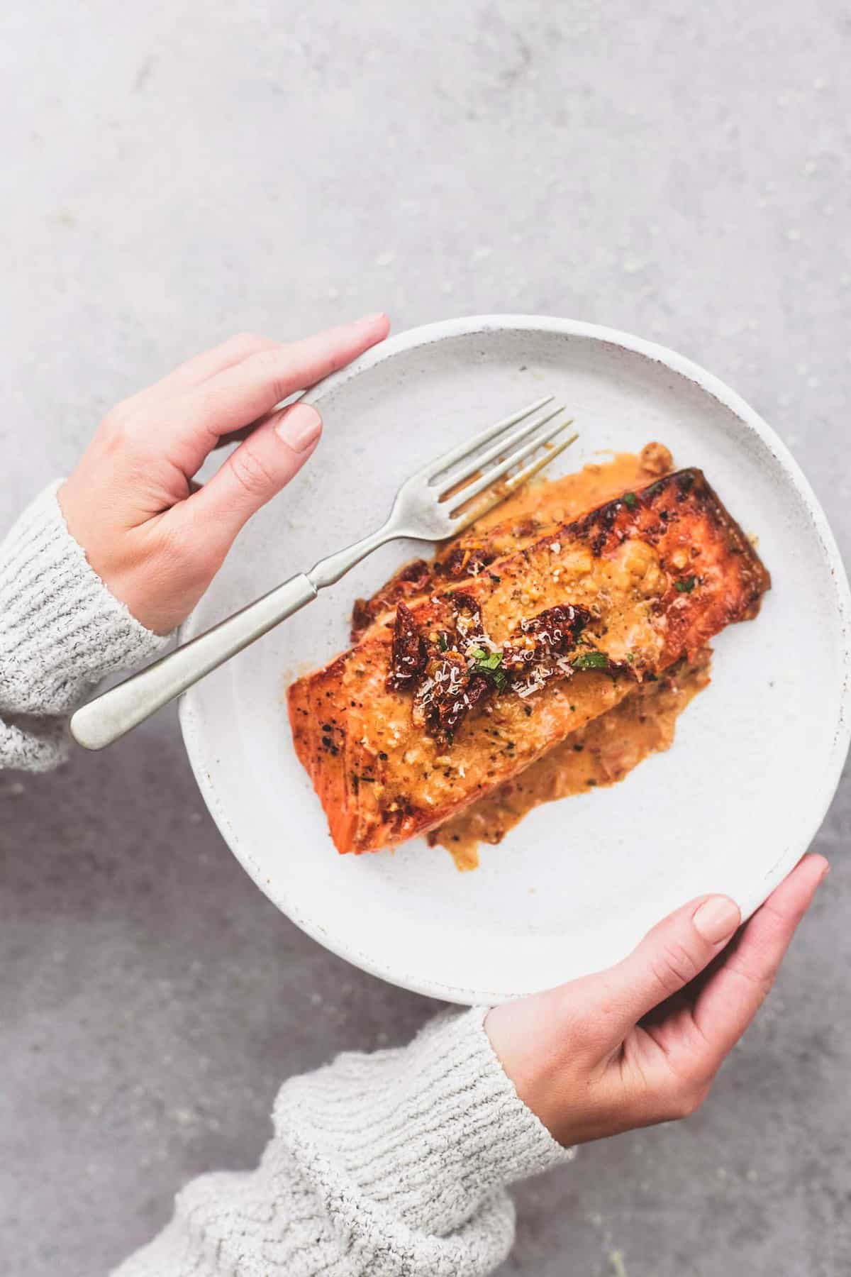 top view of hands holding a plate with salmon in creamy sun dried tomato sauce and a fork.
