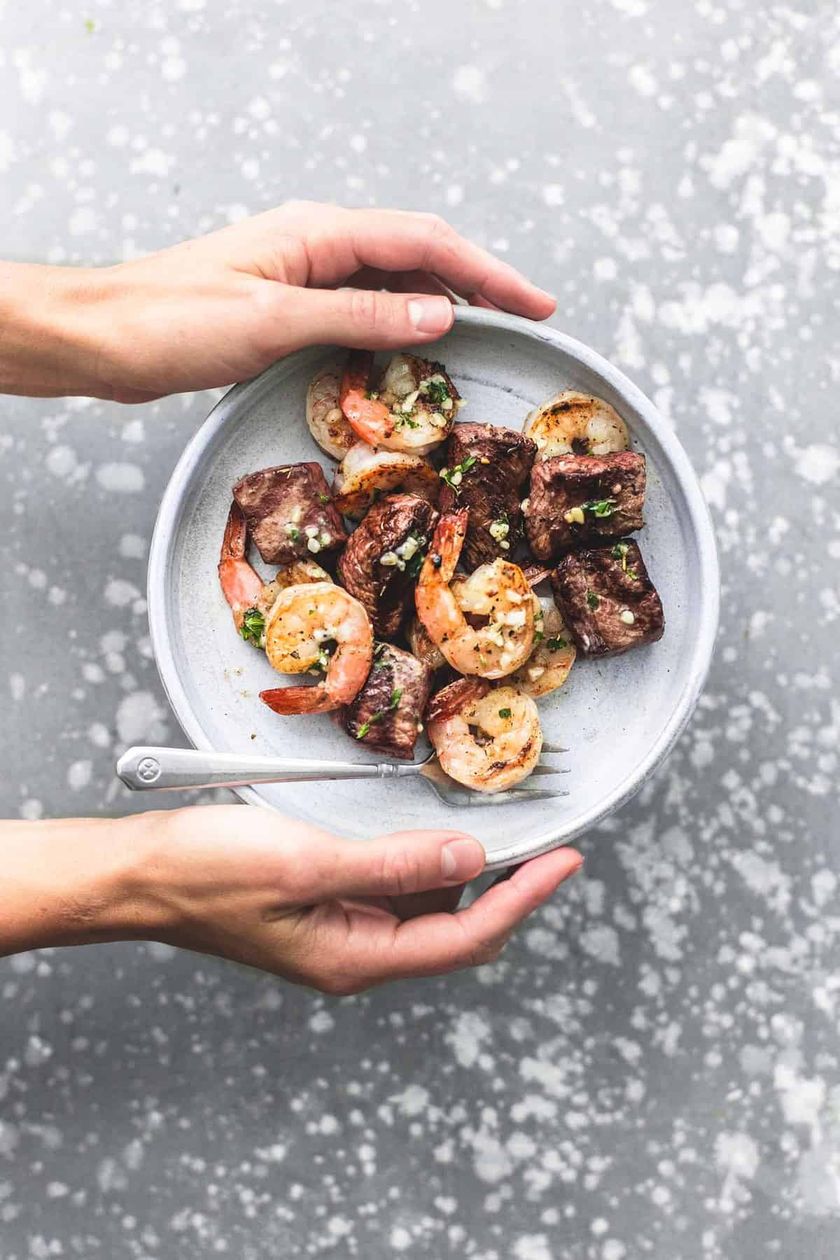 top view of a pair of hands holding deconstructed garlic butter surf and turf kabobs with a fork on a plate.