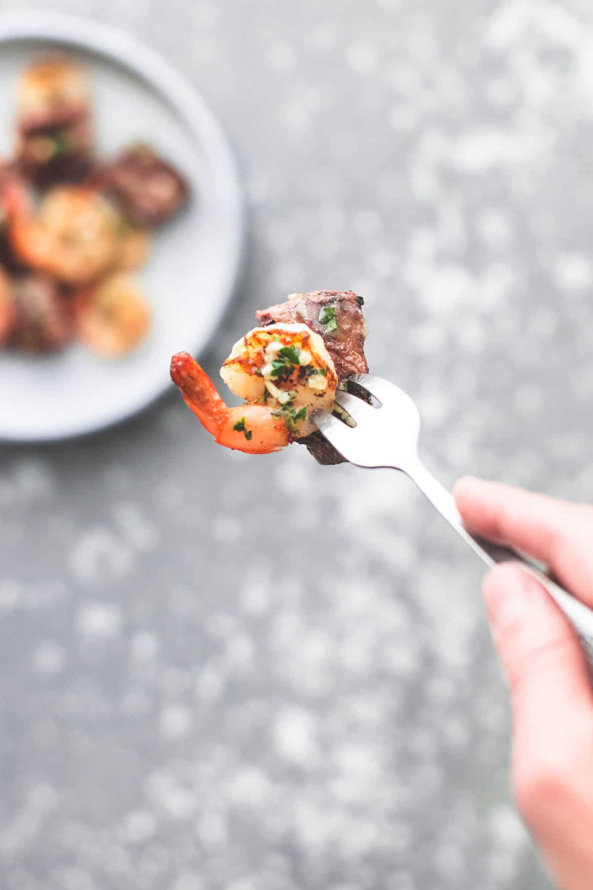 a hand holding a fork with a bite of deconstructed garlic butter surf and turf kabobs on it above a plate with more deconstructed kabobs on it.