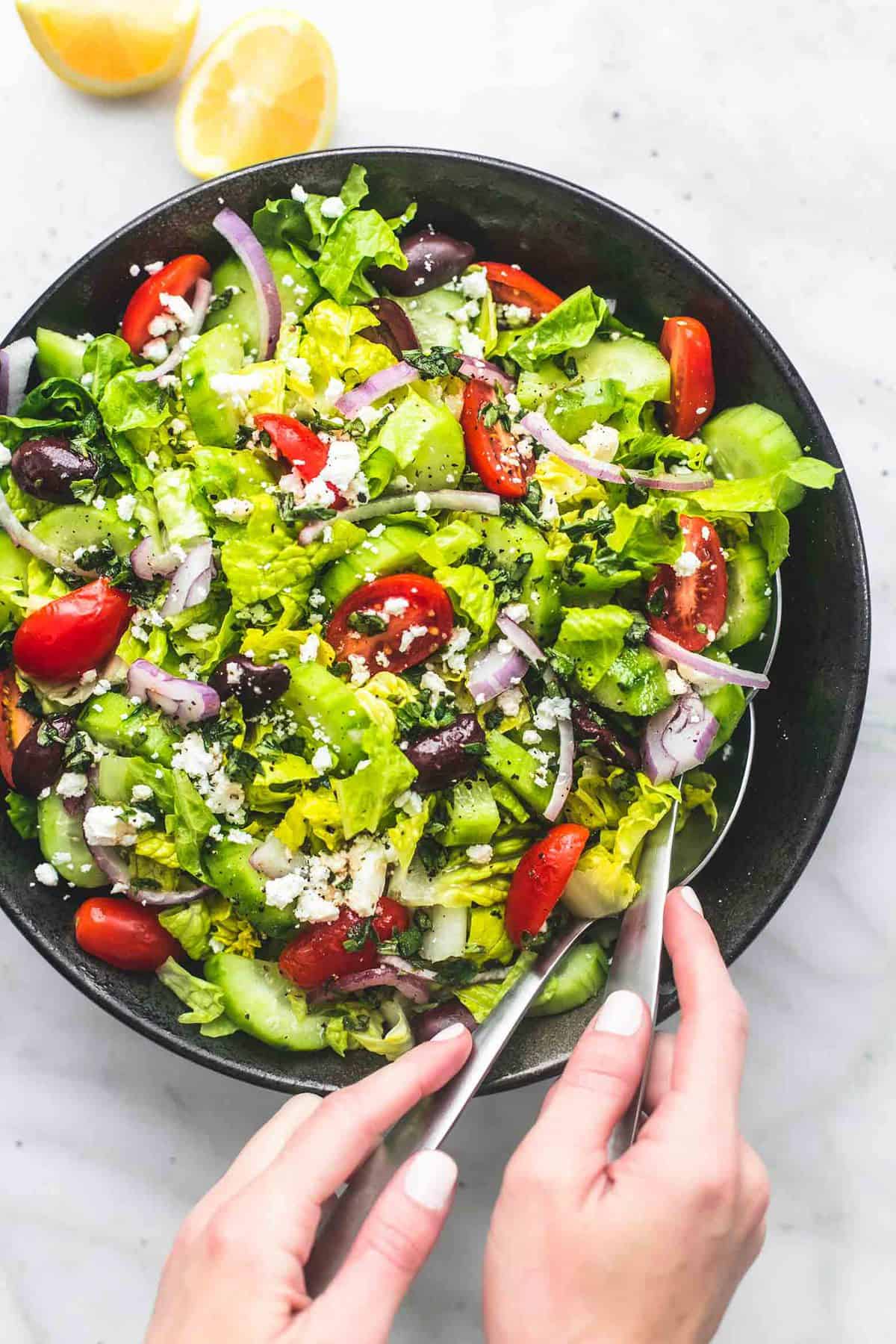 top view of Greek tossed green salad in a serving bowl with lemon slices on the side and two hands holding a serving spoon in each hand in the bowl.