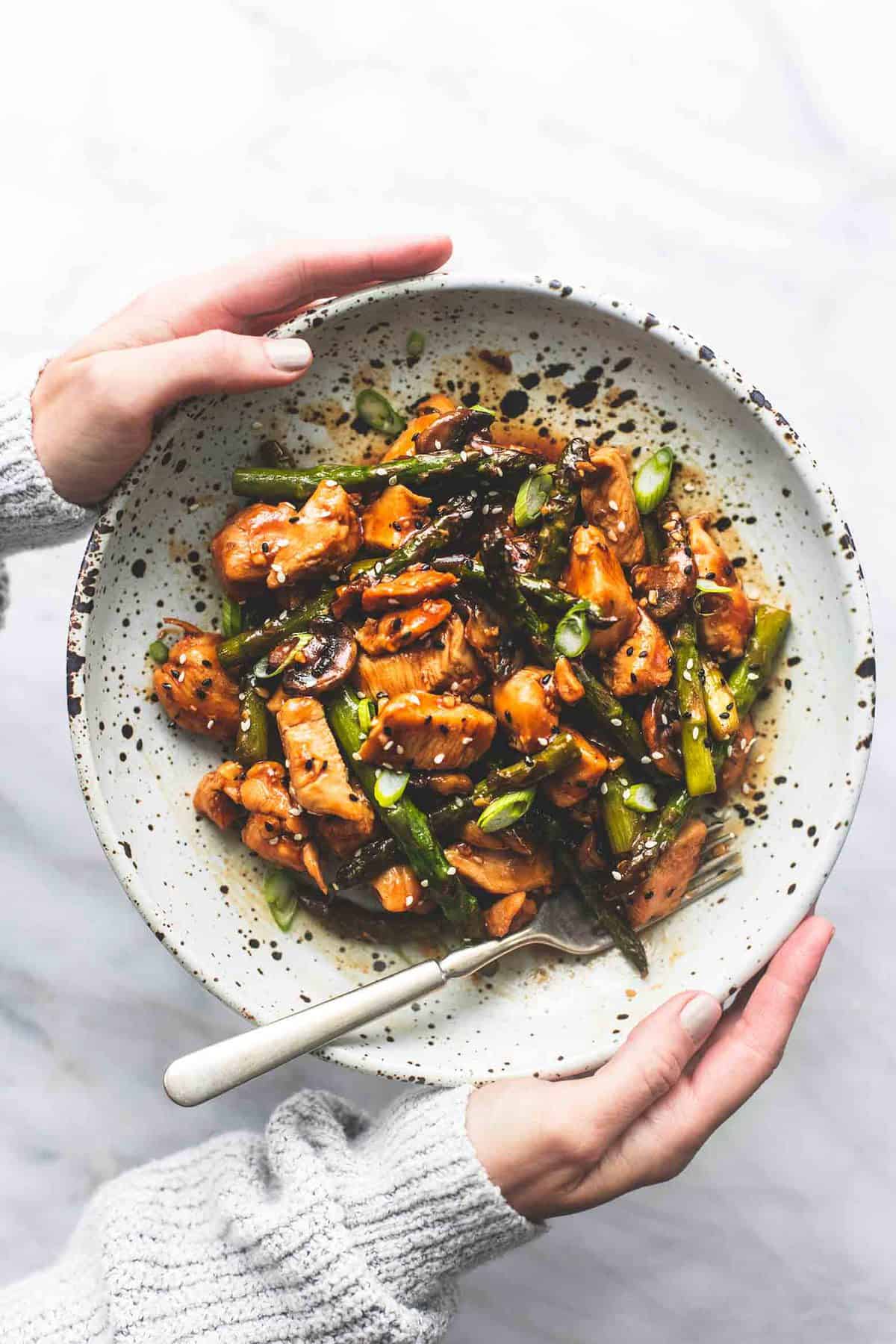 top view of a pair of hands holding a plate with chicken and asparagus stir fry and a fork on it.