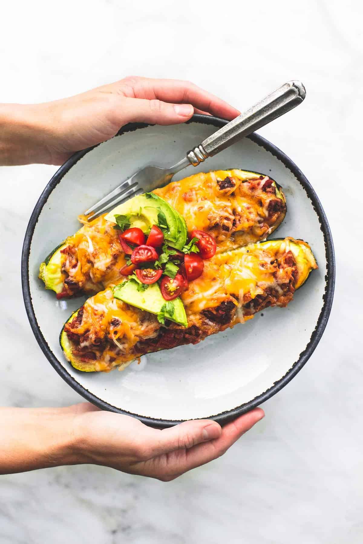 top view of a pair of hands holding a plate with taco stuffed zucchini boats and a fork on it.