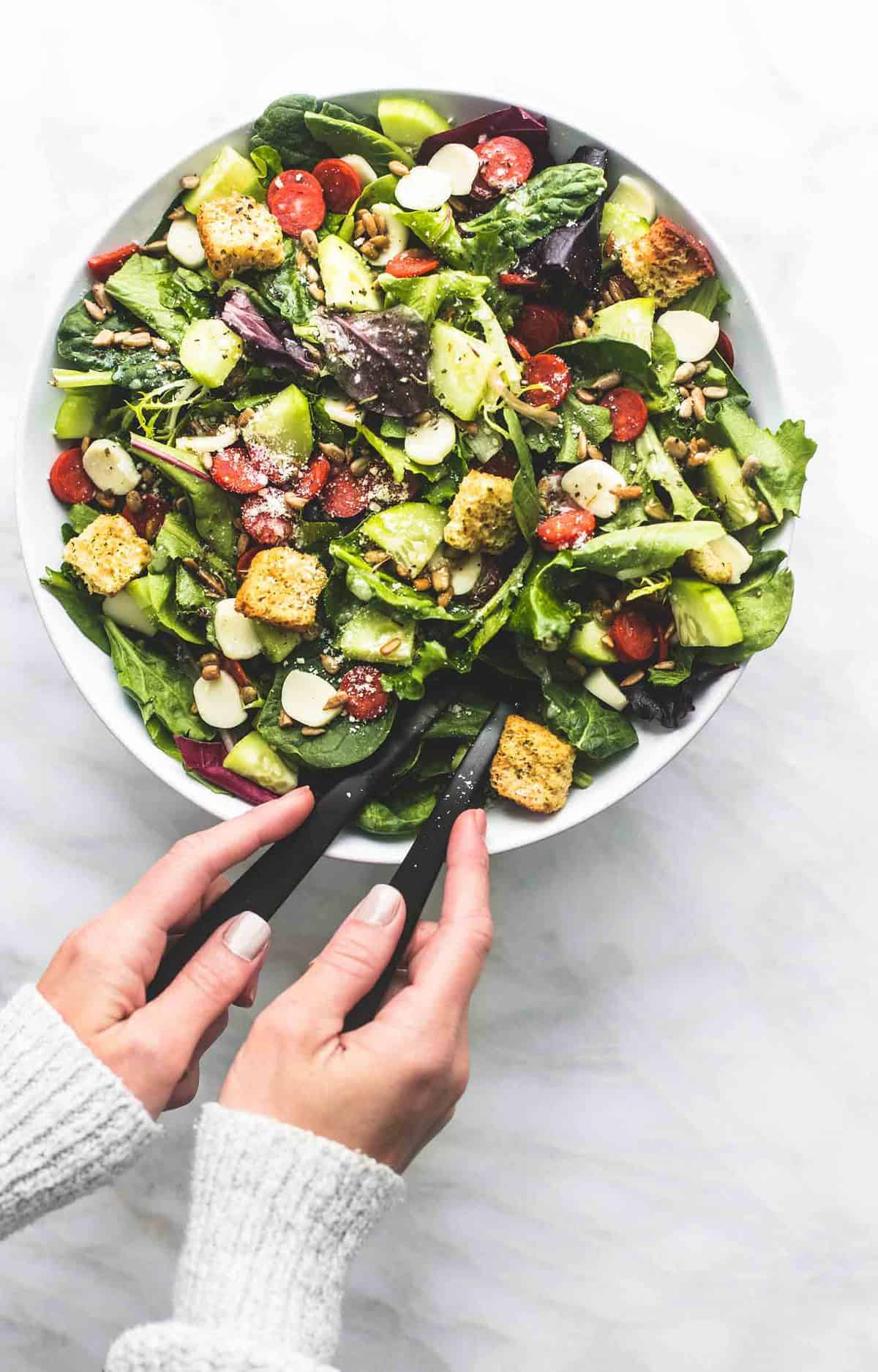 top view of Italian green salad in a bowl with two hands holding a serving spoon in each hand.