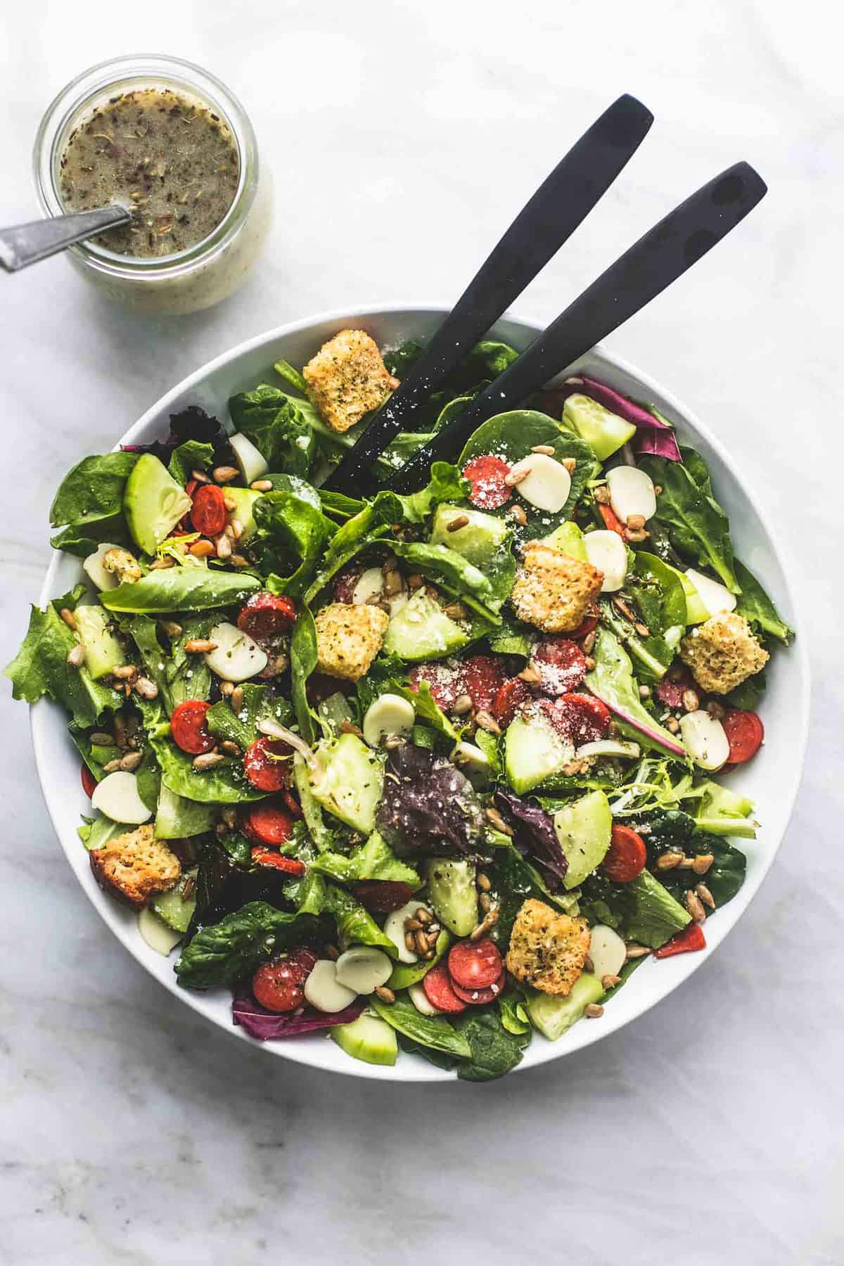 top view of Italian green salad with two serving spoons in a bowl with a jar of dressing on the side.