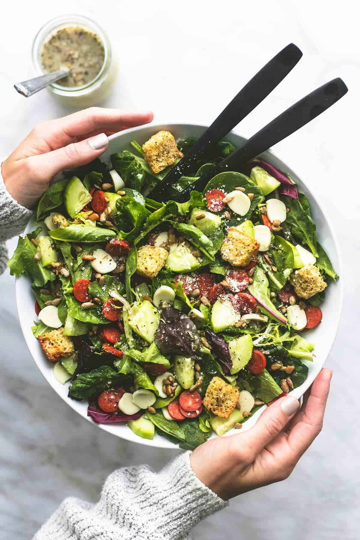 top view of a pair of hands holing a bowl of Italian green salad with two serving spoons with a jar of dressing on the side.