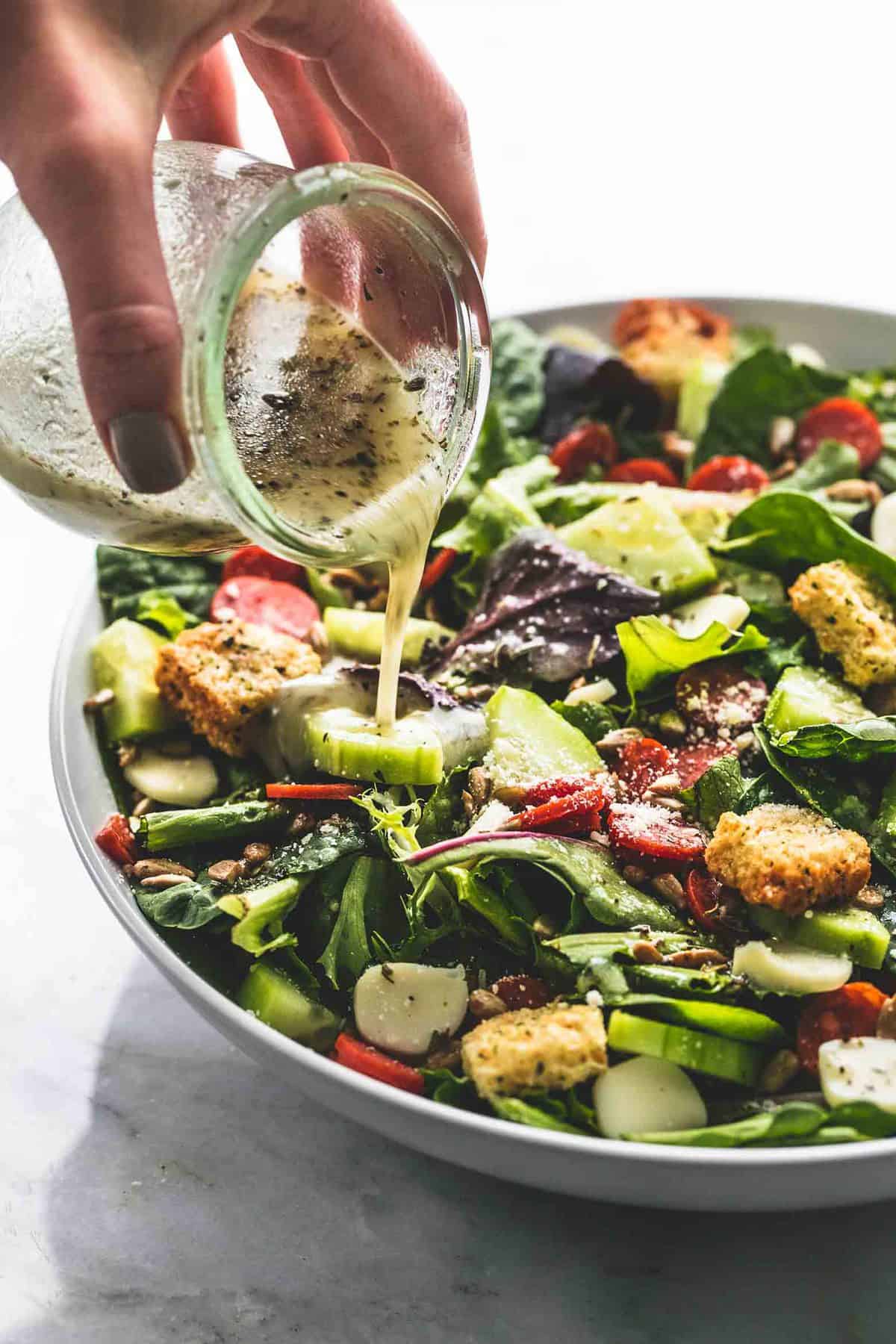 a hand pouring a jar of dressing on top of Italian green salad in a bowl.