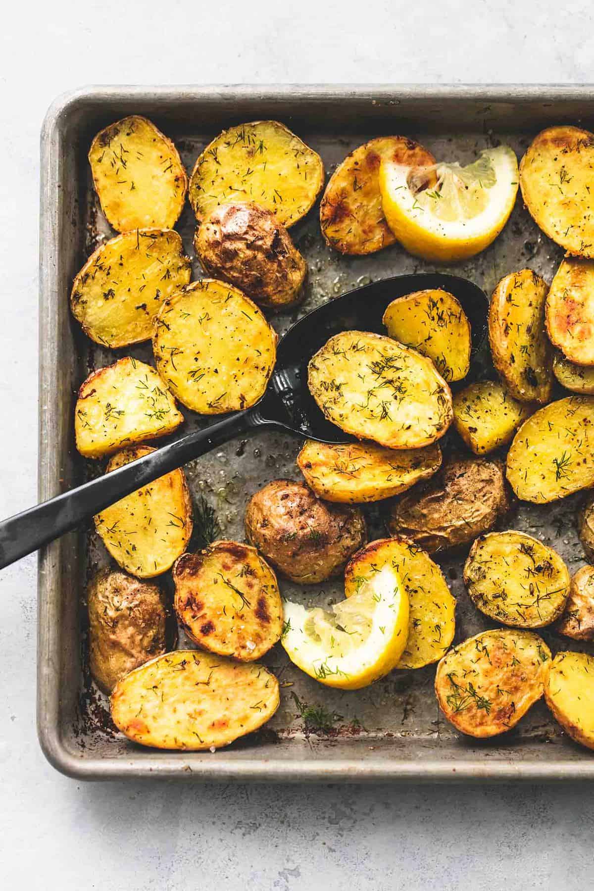 close up top view of oven roasted dill potatoes, lemon slices, and a serving spoon on a baking sheet.