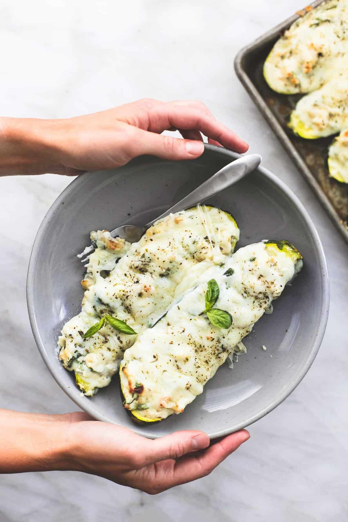 top view of a pair of hands holding a bowl with white chicken lasagna stuffed zucchini boats and a fork on it.
