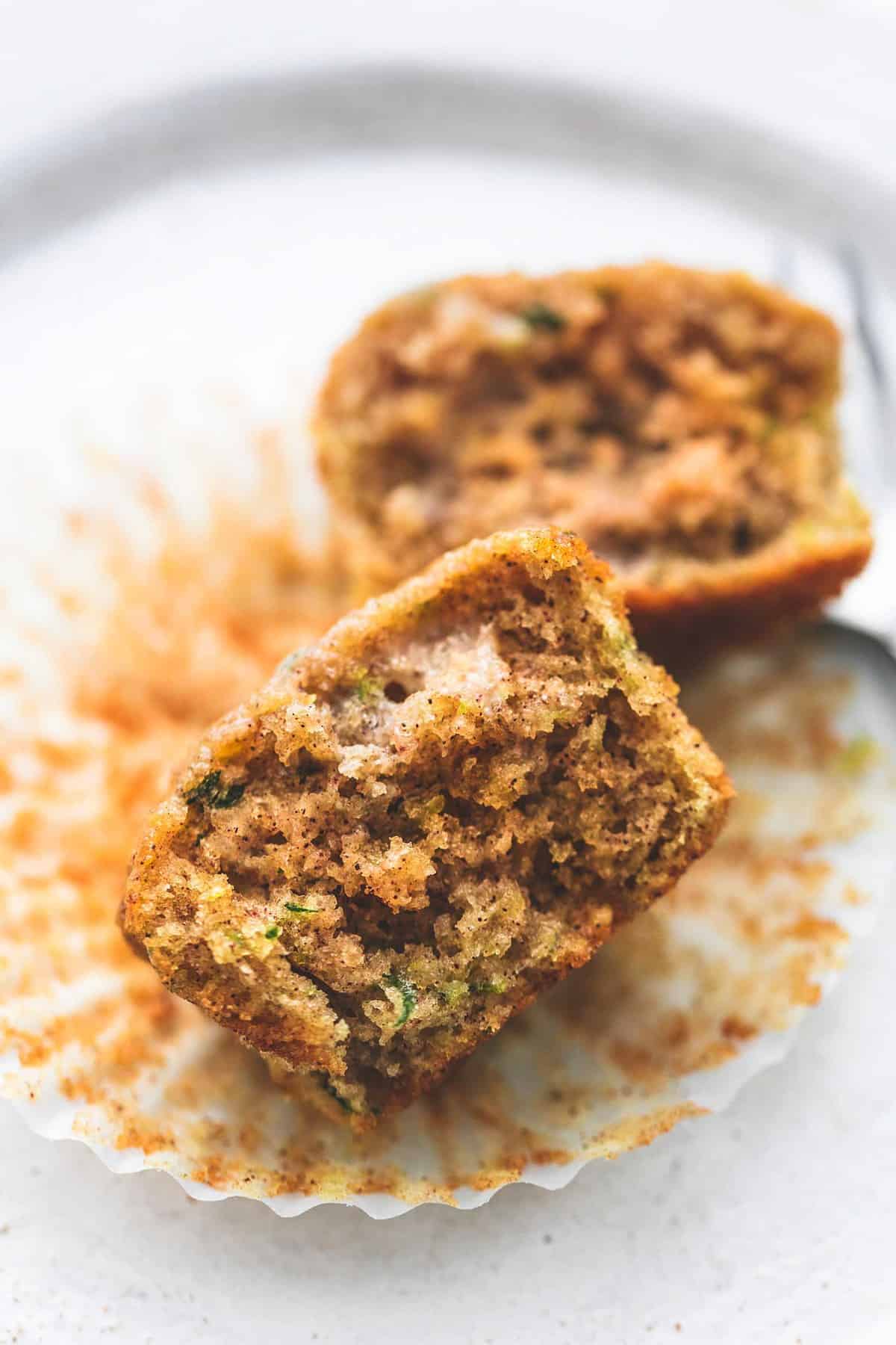 close up of a zucchini bread muffin broken in half with one half in front of the other on top of a cupcake liner with a fork on the side all on a plate.