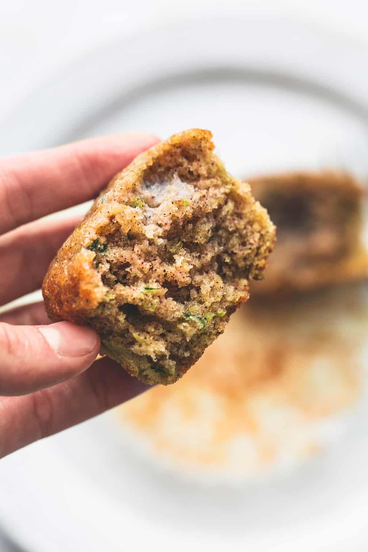 a hand holding half of a zucchini bread muffin above a plate with the other half on it faded underneath.