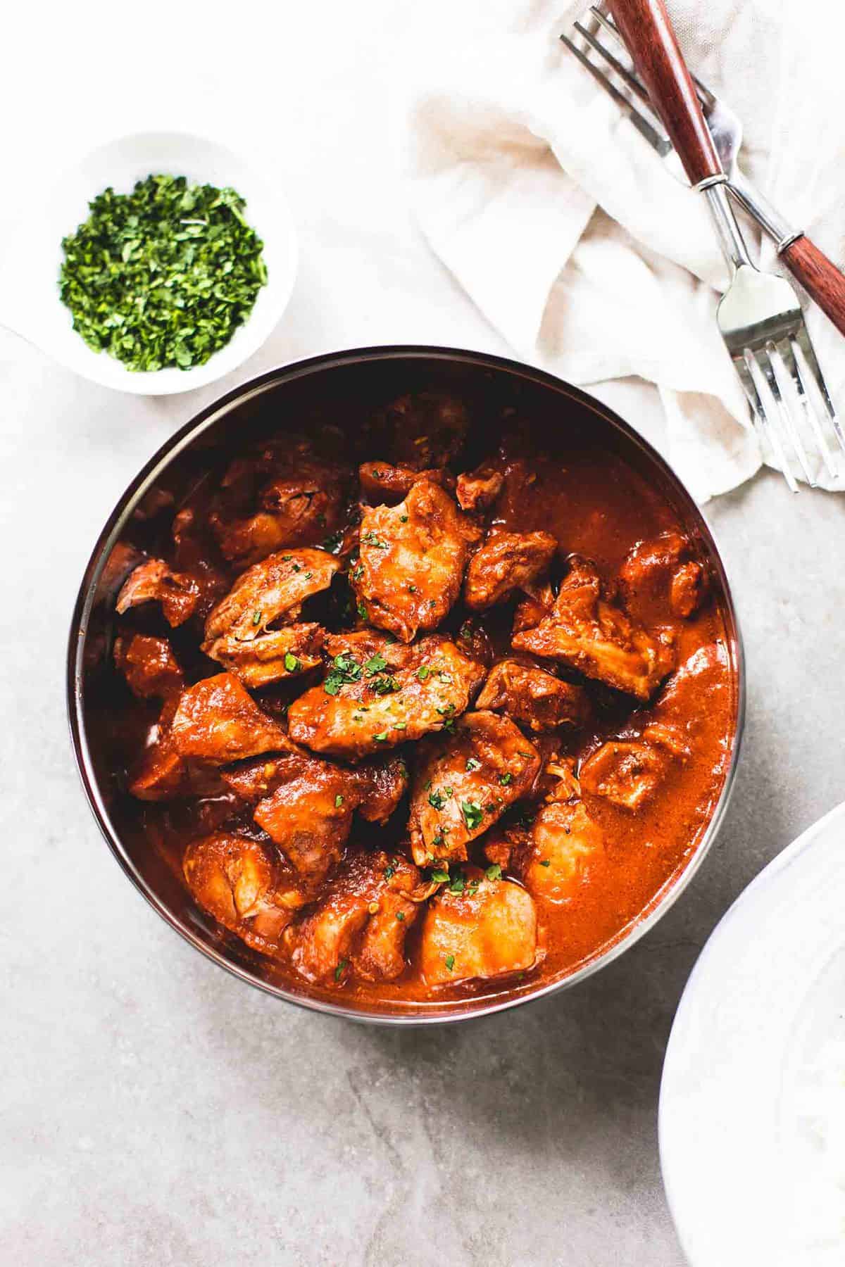 top view of instant pot chicken tikka masala in a pan with a bowl of cilantro and two forks on a cloth napkin on the side.