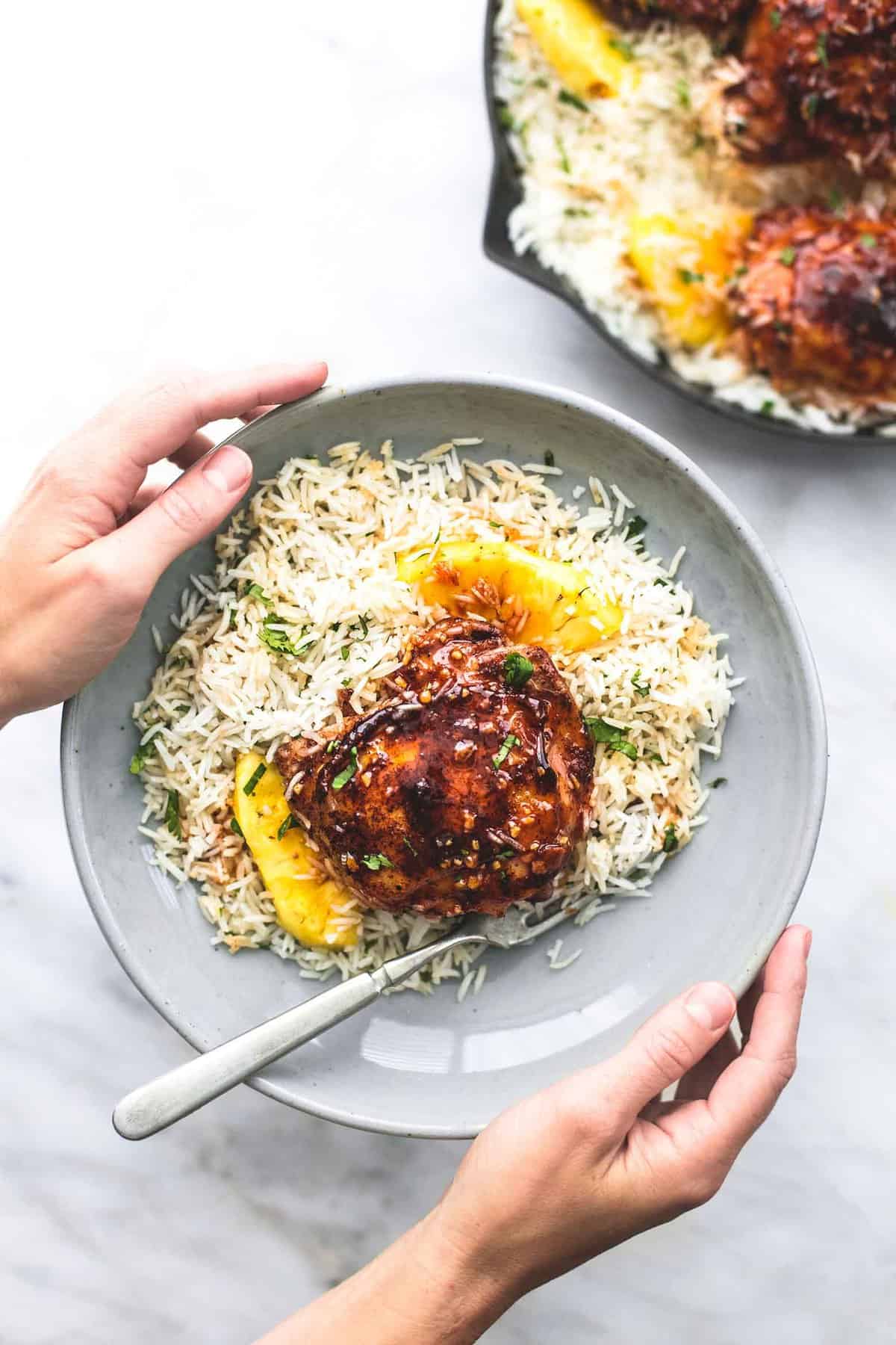 top view of a pair of hands holding a plate with Island glazed chicken and coconut rice and a fork on it with a pan of more rice and chicken on the side.