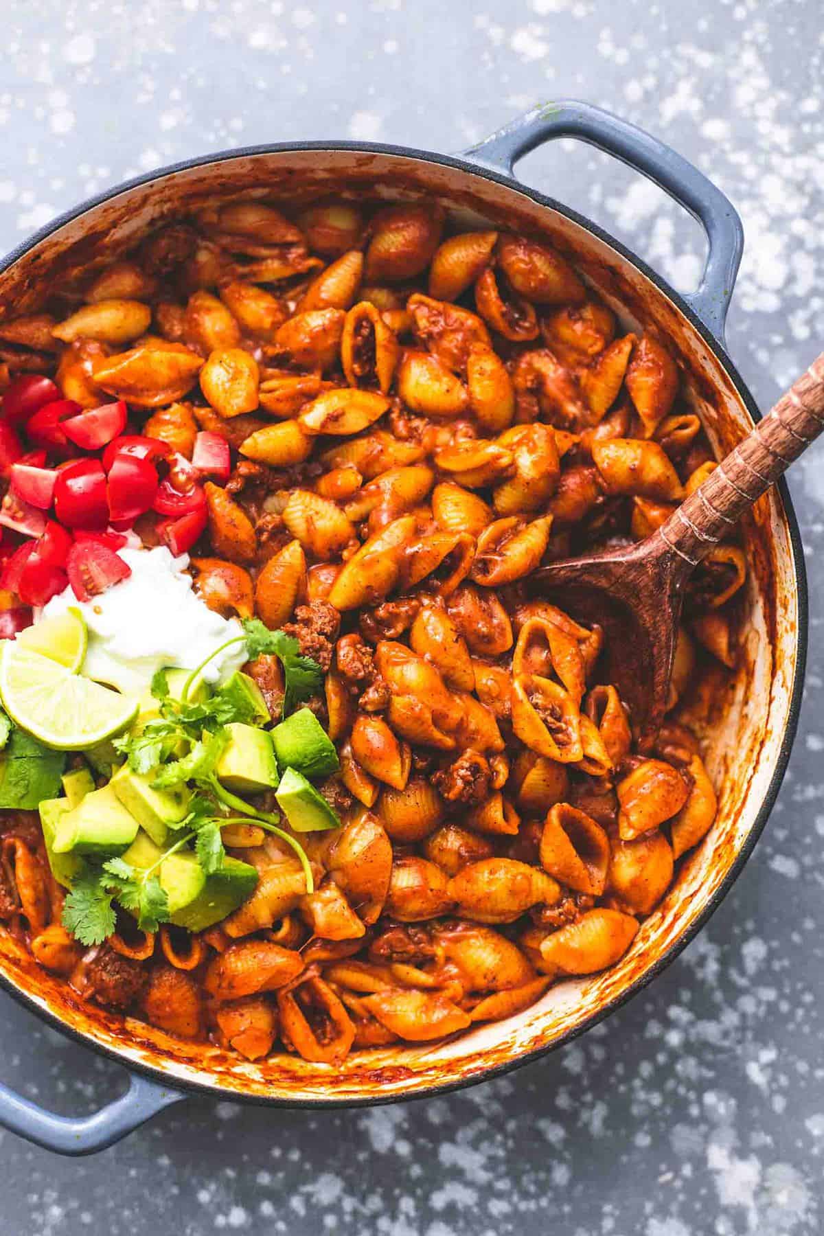 top view of beefy enchilada shells with taco toppings and a wooden serving spoon on the sides all in a pan.