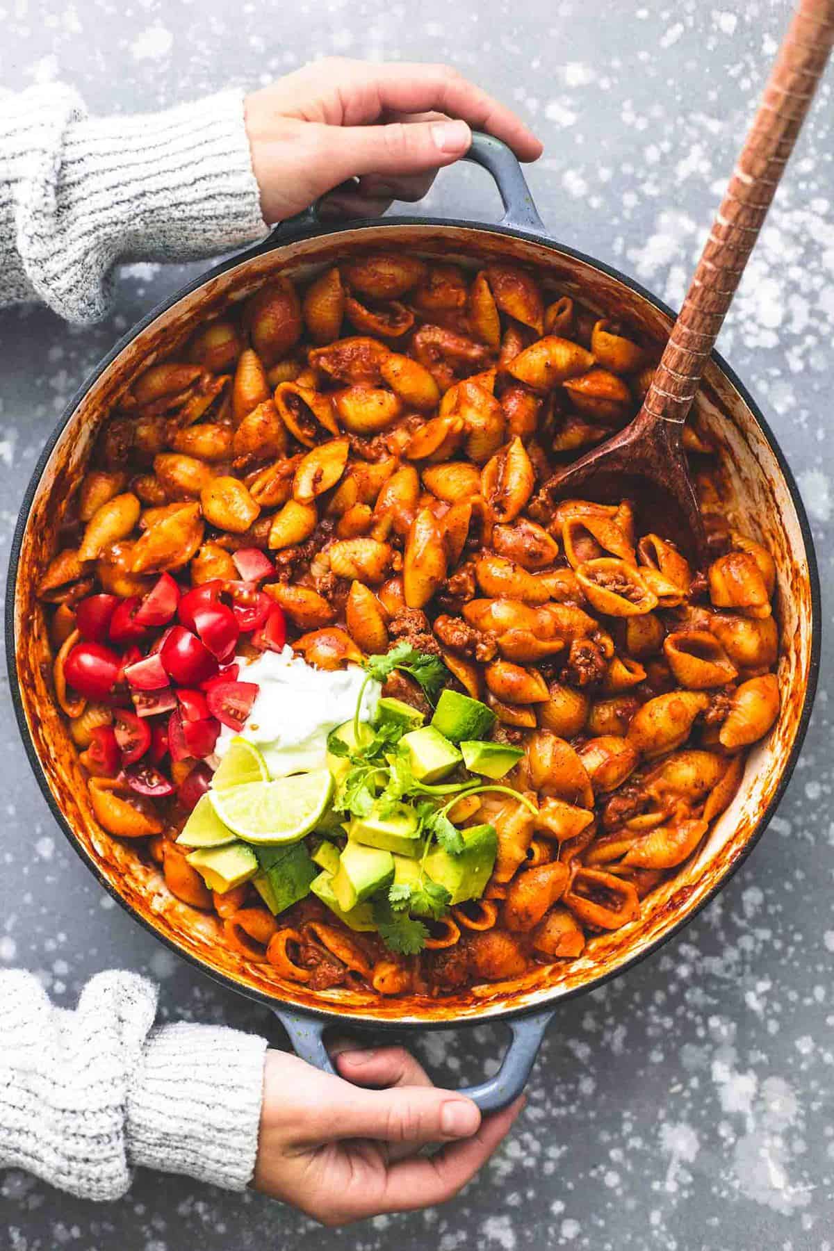 top view of a pair of hands holding each handle on the side of a pan of beefy enchilada shells with taco toppings and a wooden spoon on the sides.