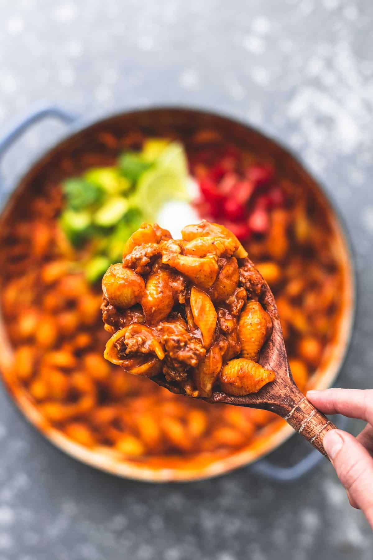 top view of a hand holding a scoop of beefy enchilada shells with a wooden serving spoon above a pan of shells.