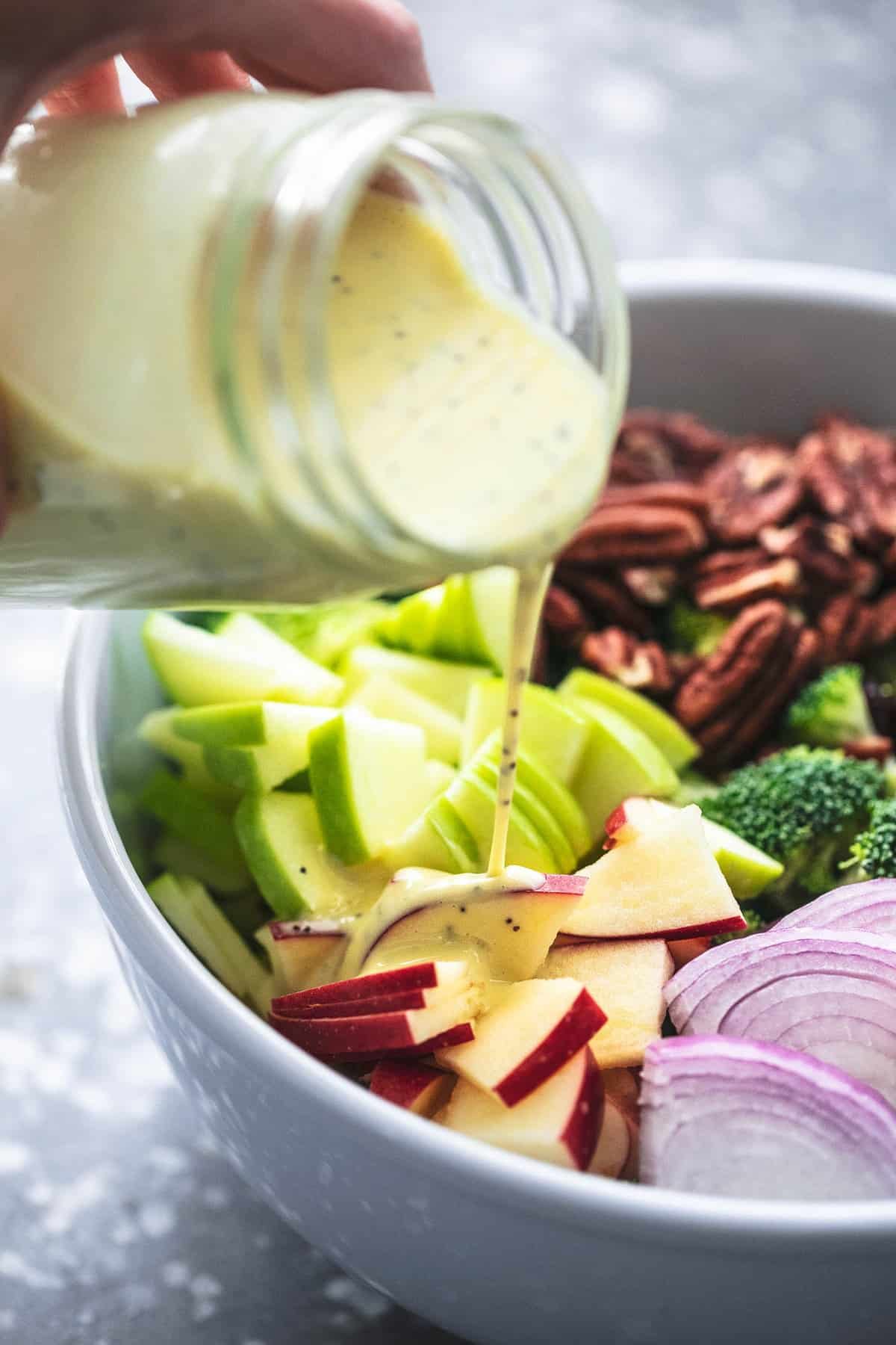 a hand pouring a jar of poppyseed dressing on top of broccoli apple salad not yet tossed.