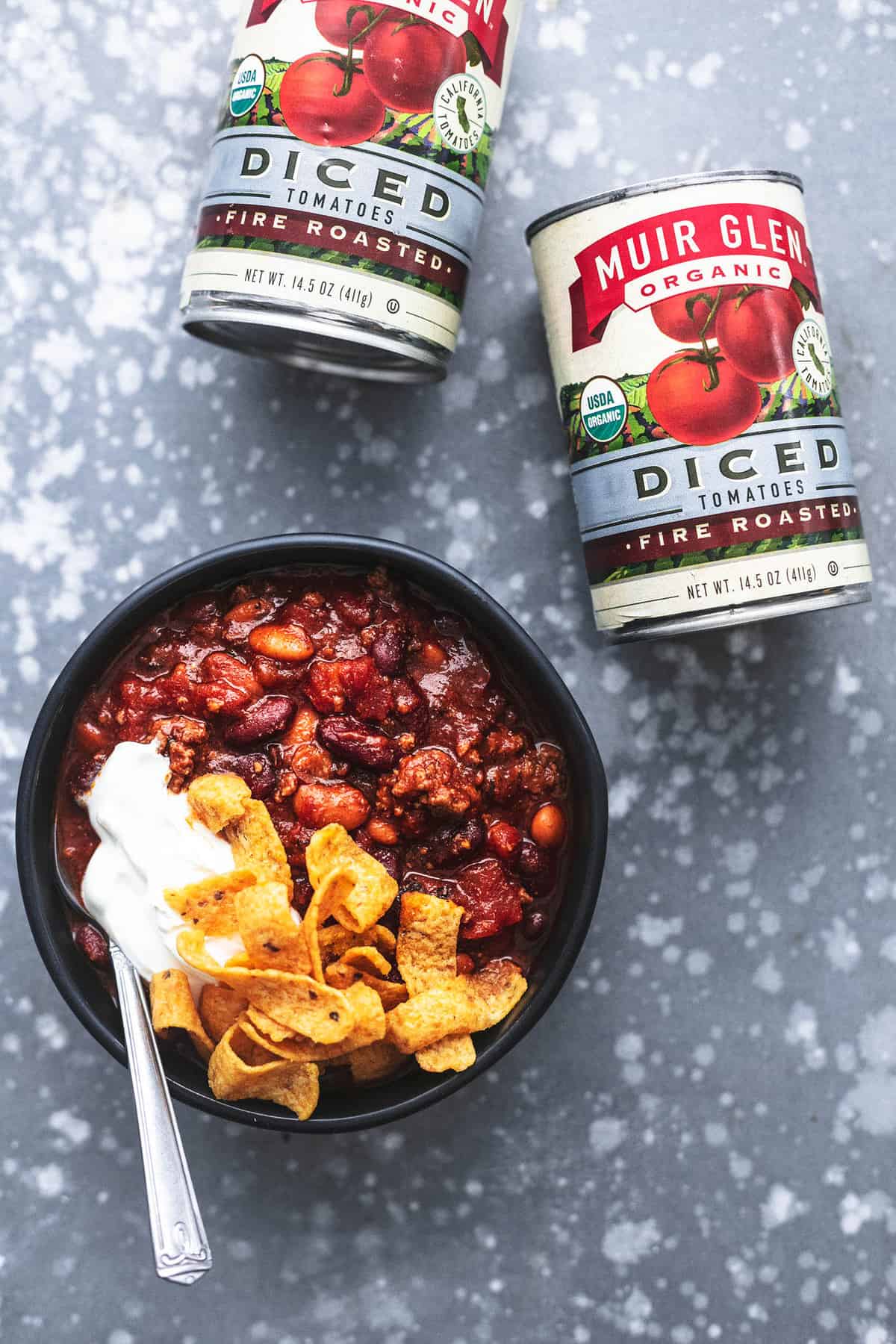 top view of cans of Muir Glen Organic diced tomatoes with a bowl of chili with Fritos, sour cream and a spoon.