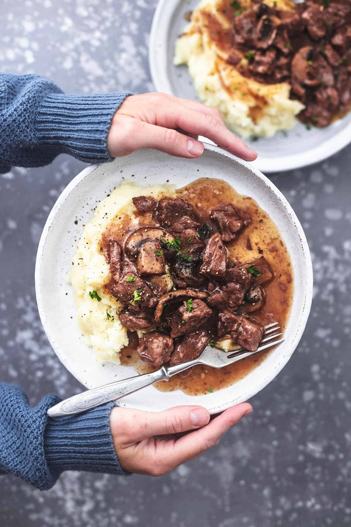 top view of a pair of hands holding a plate with instant pot beef tips a fork and mashed potatoes on it with another plate on the side.