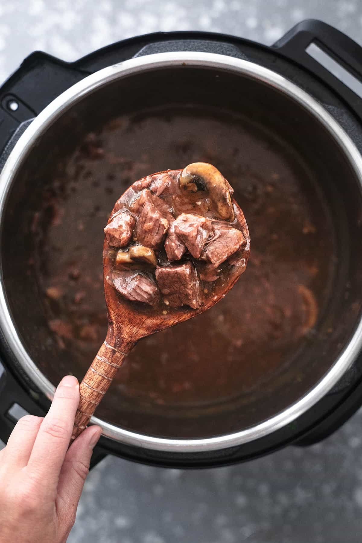 top view of a hand holding a scoop of instant pot beef tips with a wooden serving spoon above an instant pot of more beef tips.