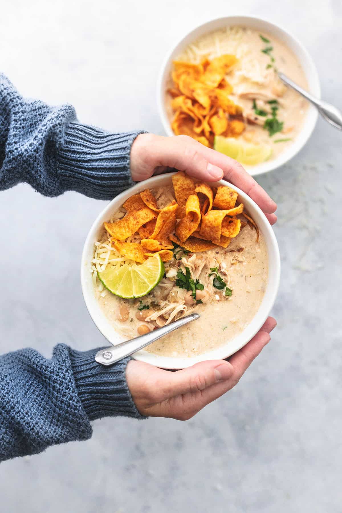 top view of a pair of hands holding instant pot white chicken chili with Fritos, a lime slice and a spoon in a bowl with another bowl on the side.