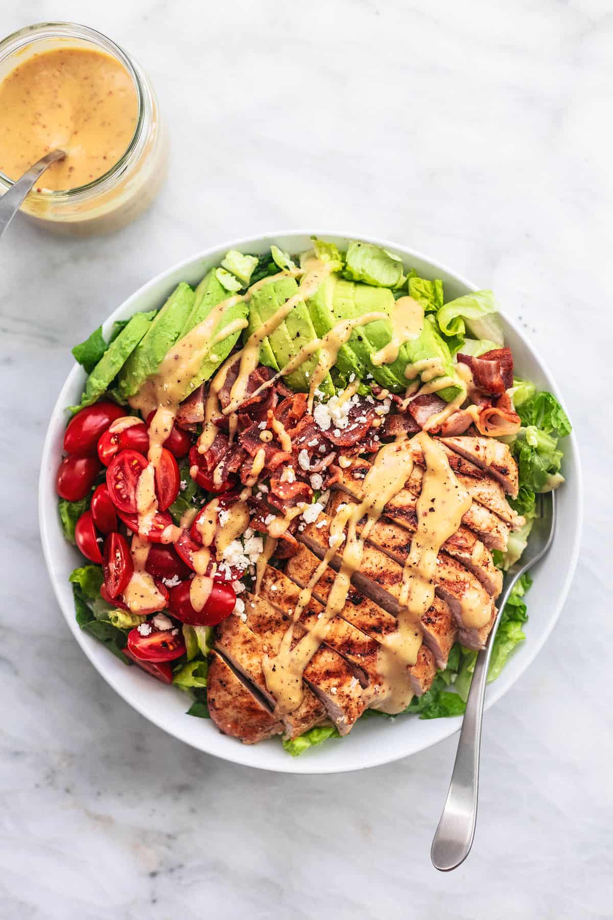 top view of chicken avocado salad with a fork on a plate with dressing with a spoon in a glass on the side.