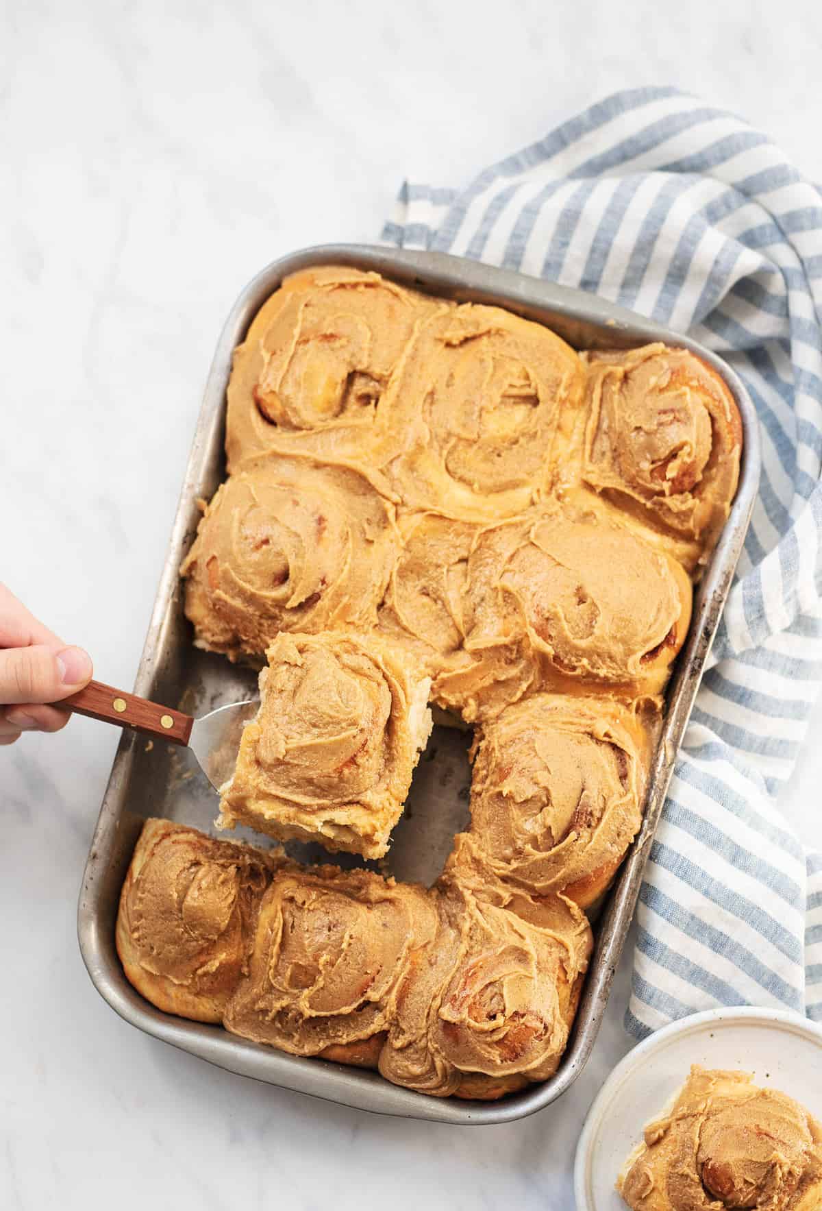 top view of a hand lifting a cinnamon roll with brown sugar frosting with a spatula from a baking pan.