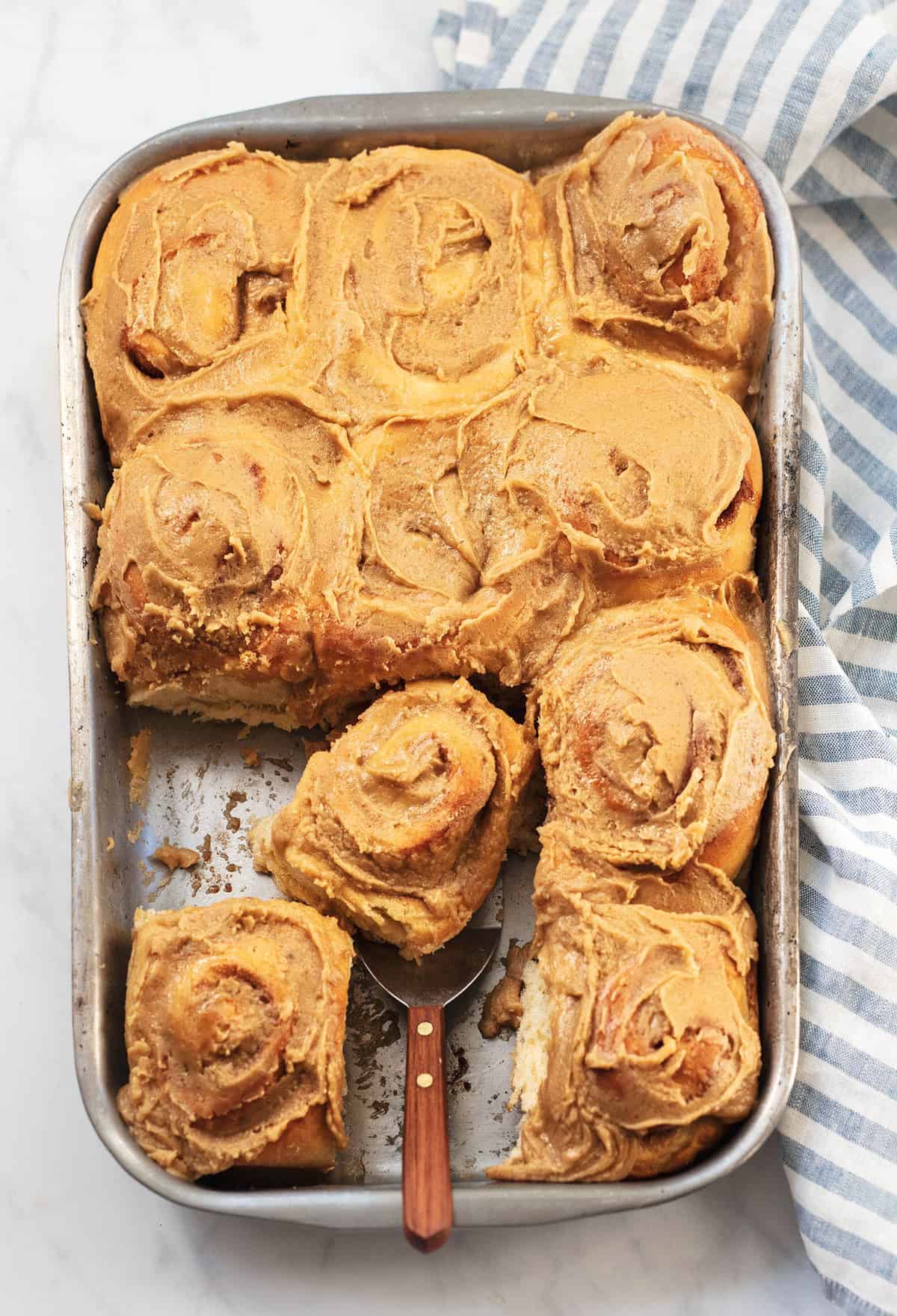 top view of cinnamon rolls with brown sugar frosting with some missing and a roll on a spatula in a baking pan.