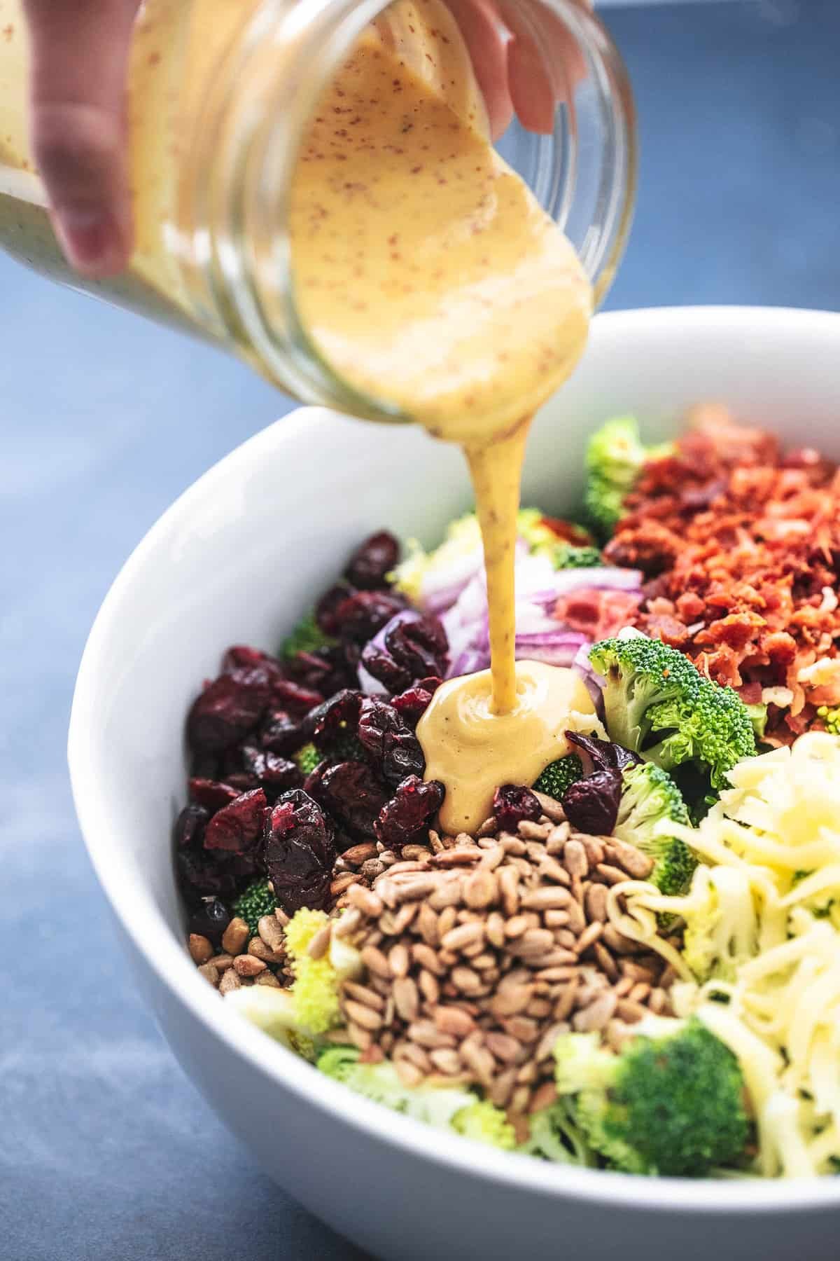 close up of a hand pouring a jar of dressing on top of broccoli salad not yet mixed in a serving bowl.