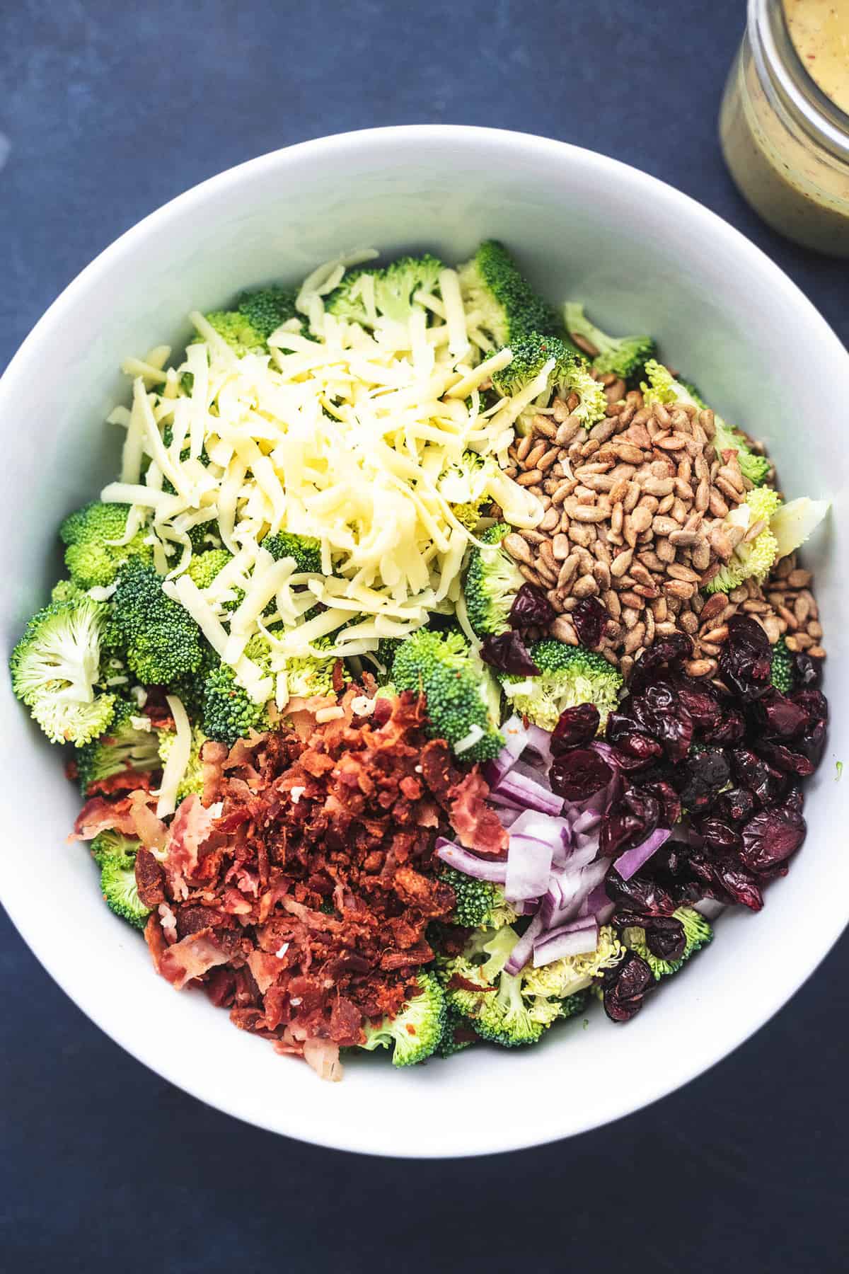 top view of broccoli salad ingredients not mixed in a serving bowl with a jar of dressing on the side.