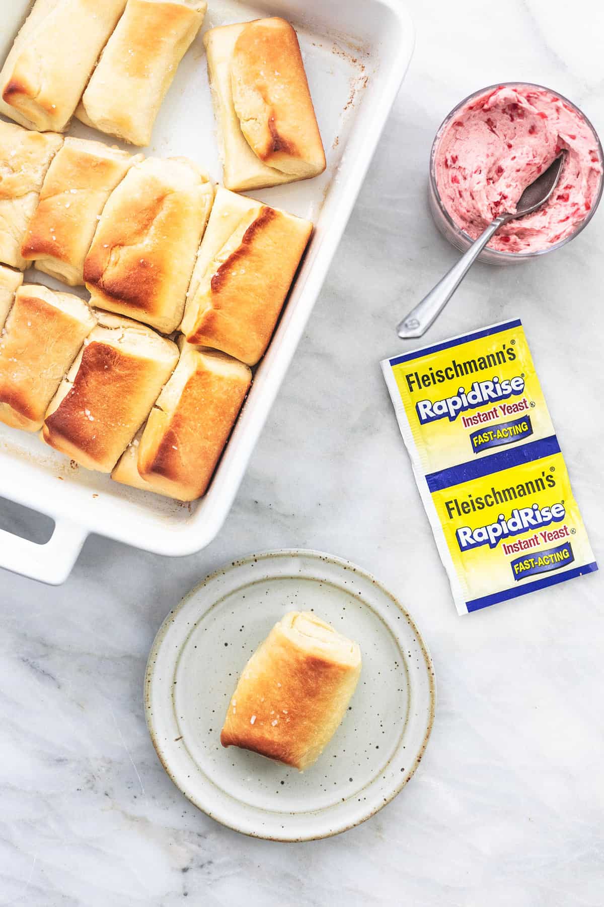 top view of a homemade Parker House roll on a plate with a baking pan with more rolls, RapidRise yeast packets. and a small glass bowl of raspberry almond butter with a spoon on the side.