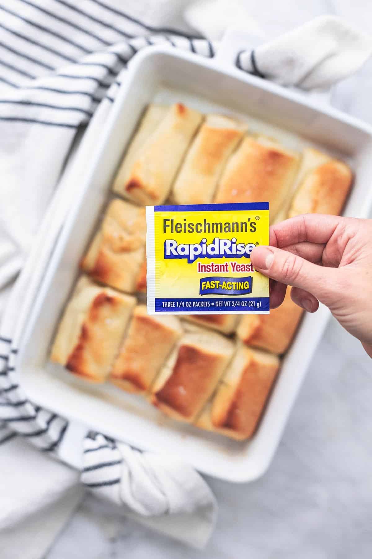 a hand holding a RapidRise yeast packet above a baking pan of homemade Parker House rolls.