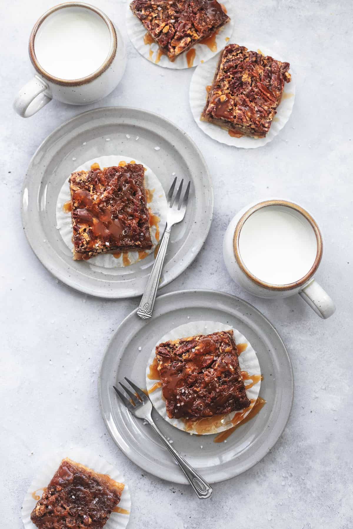 top view of two plates with forks and salted caramel pecan pie bars on them and two mugs of milk and more bars on the side.