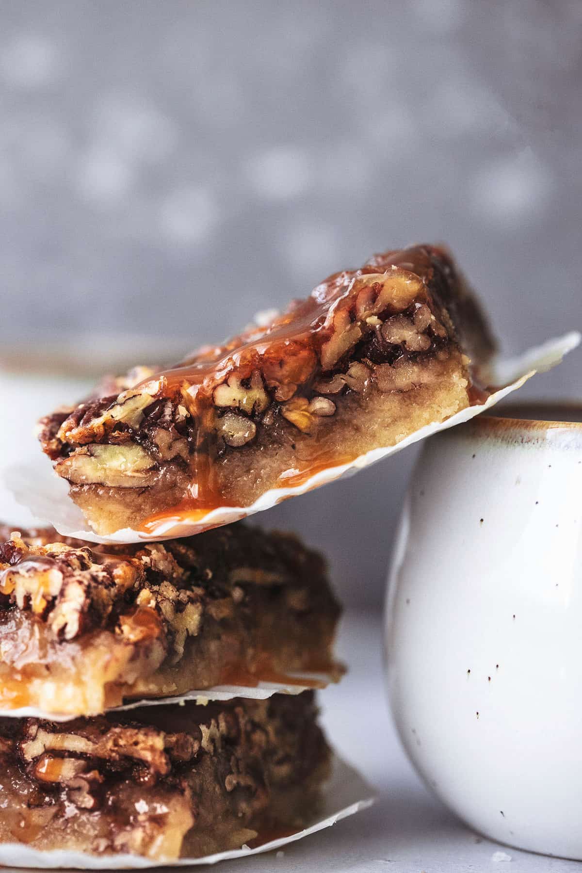 stacked salted caramel pecan pie bars with cupcake liners in between each bar and the top bar leaning on the rest of the bars and on a mug.