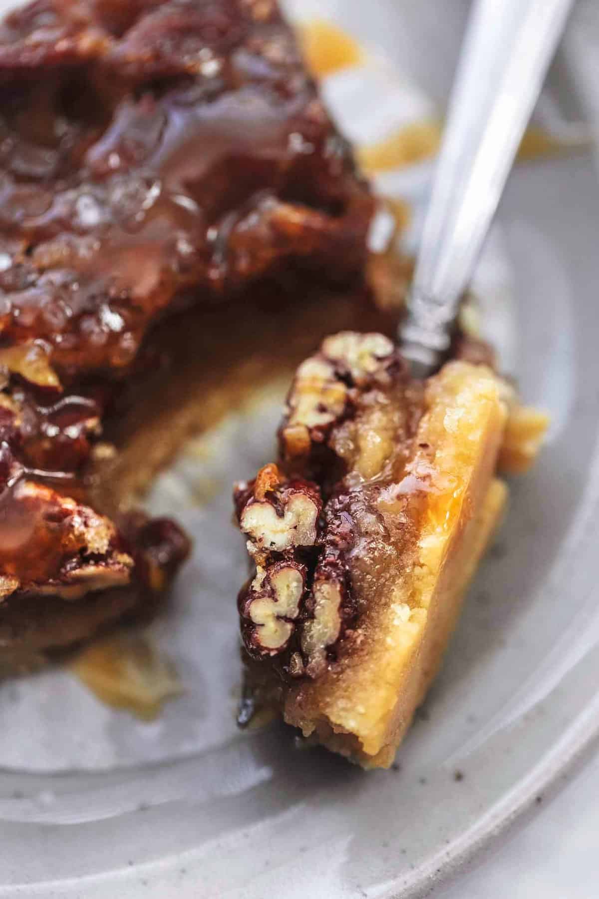 close up of a bite of a salted caramel pecan pie bar on a fork with the rest of the bar on the side both on a plate.