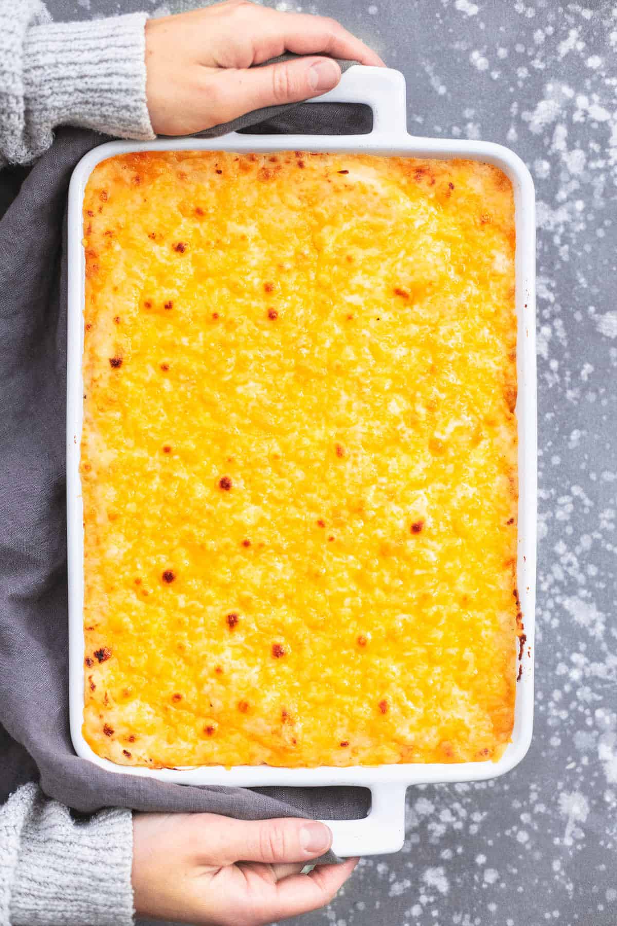 top view of hands holding the handles of a baking pan of cheesy mashed potatoes casserole.