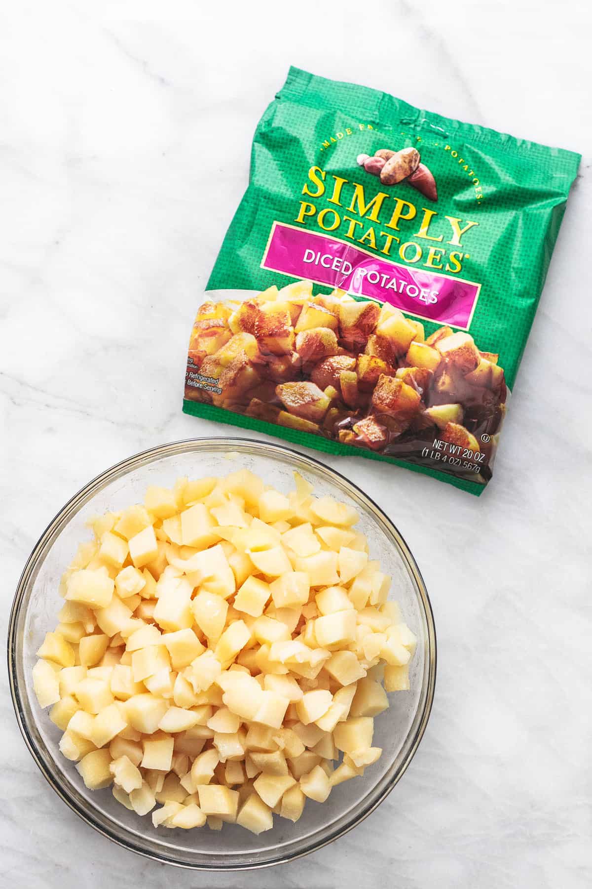 top view of raw potatoes cut up into small cubes in a glass bowl with a package of Simply Potatoes on the side.