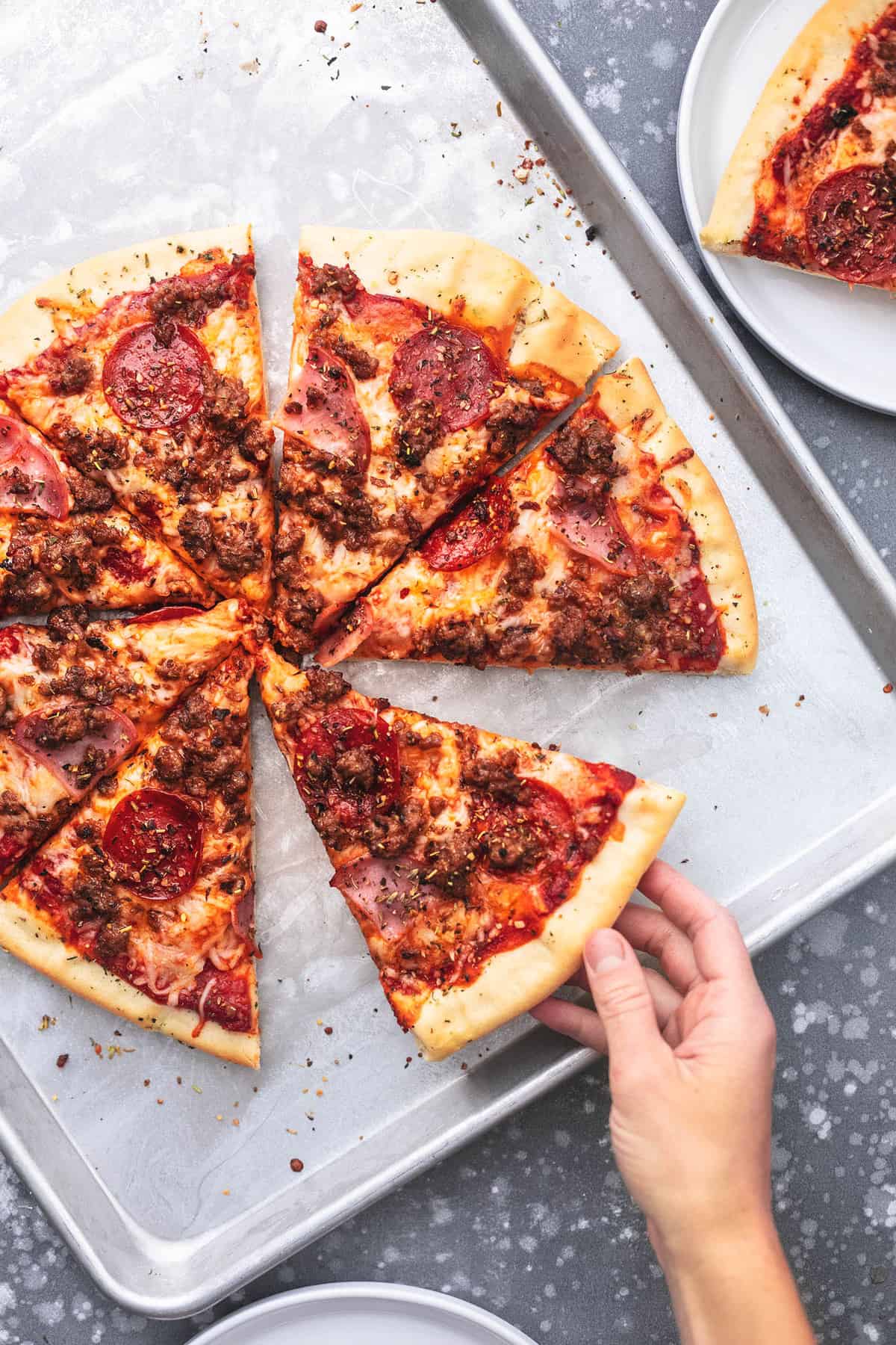 top view of meat lovers pizza cut into slices on a baking sheet with a hand taking one of the slices.