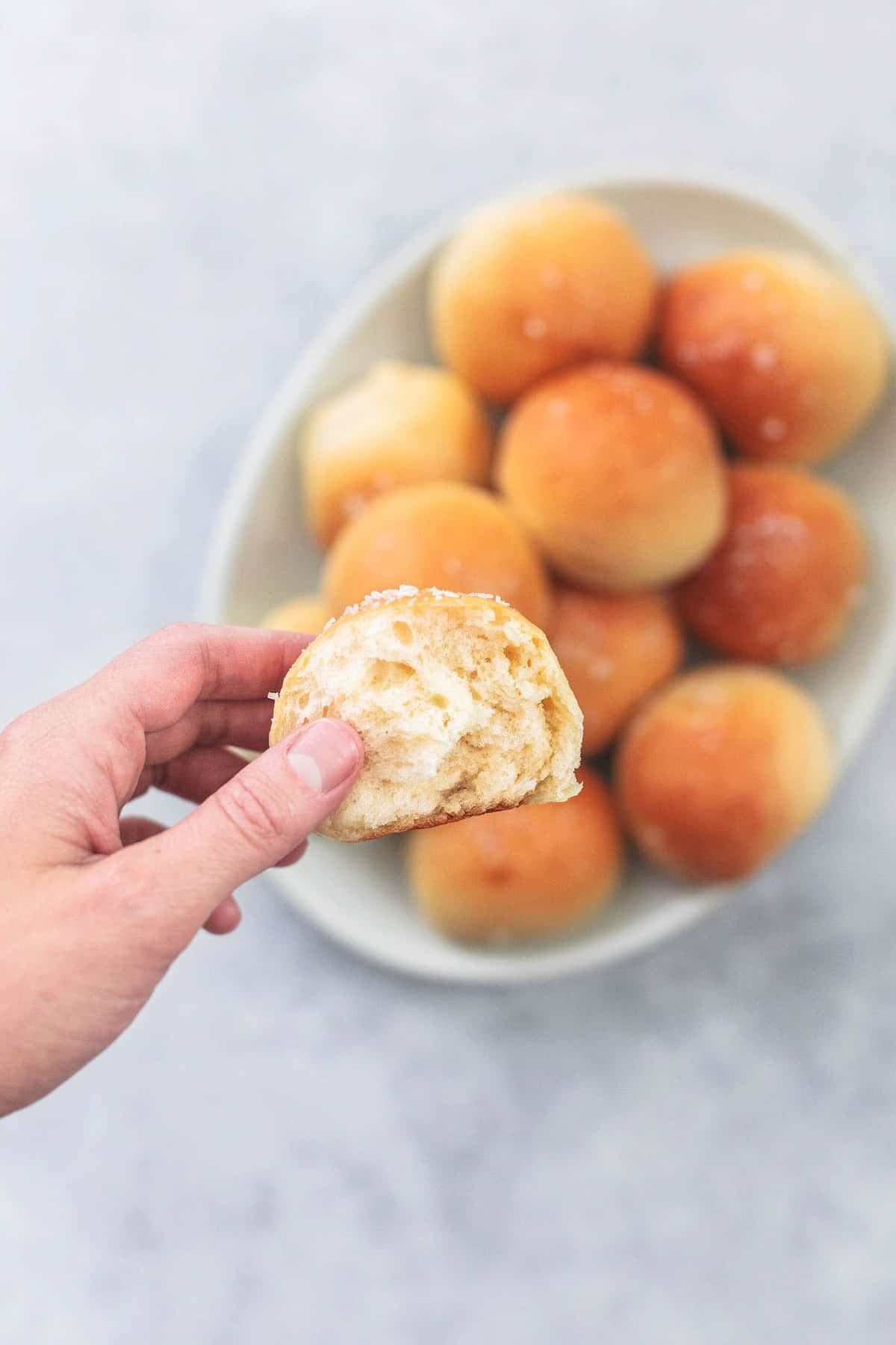 a hand holding up half of a potato roll above a plate of rolls.